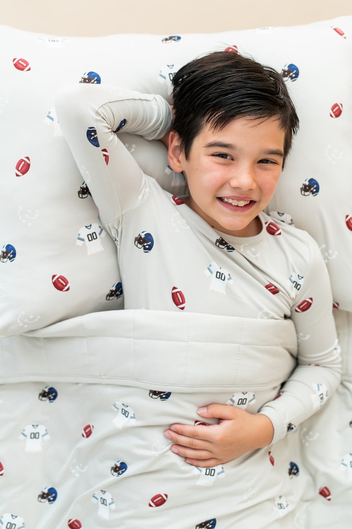 Boy lying on Standard Pillowcase and sheets in Vintage Football print, wearing a similar print Long sleeve pajama set under a similar print youth blanket