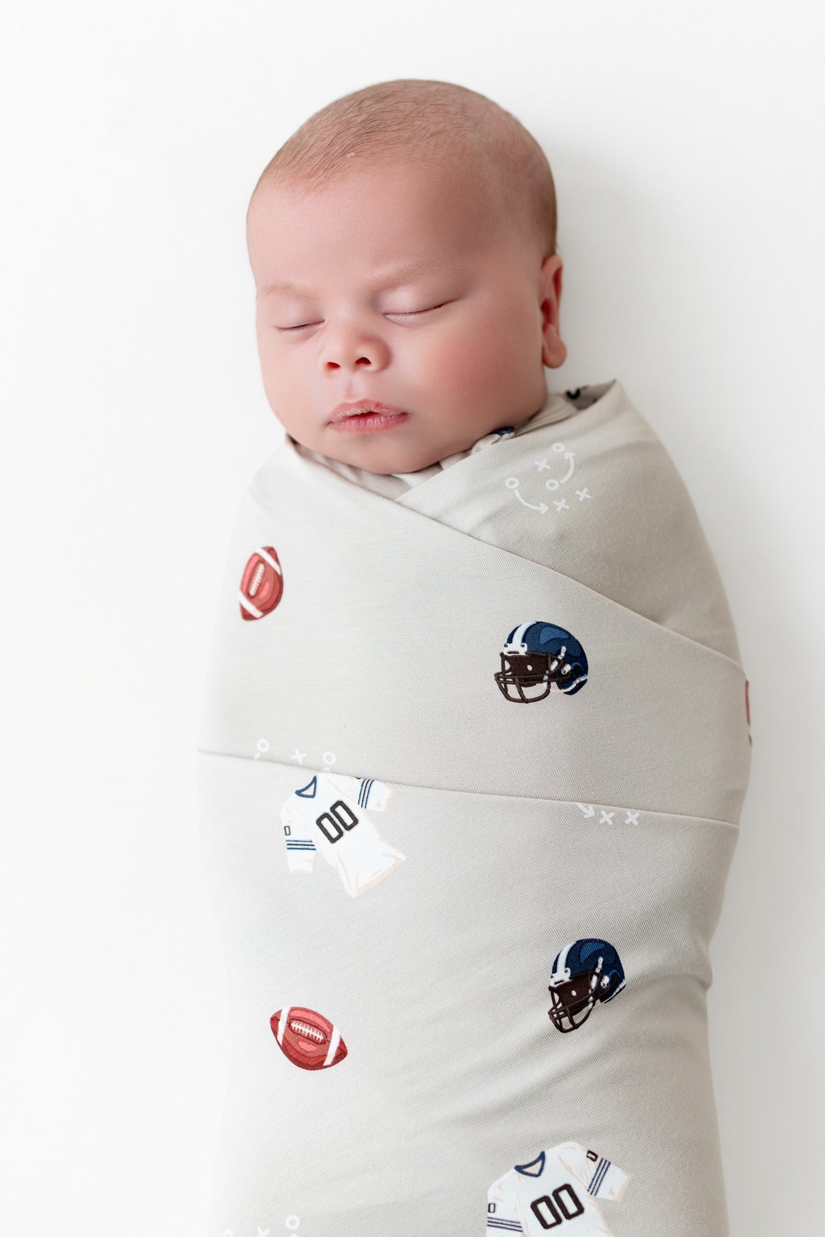 A sleeping newborn baby swaddled in a light beige fabric wrap with a pattern of footballs and football helmets, photographed against a clean white background.