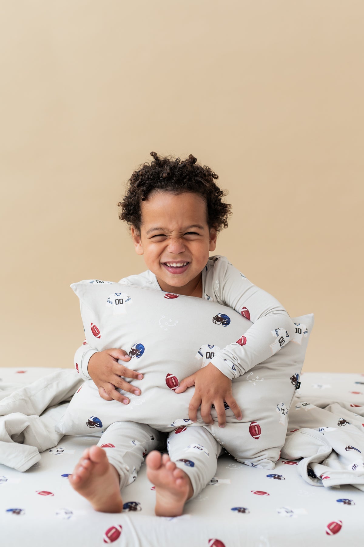 Boy holding Toddler Pillowcase in Vintage Football print sitting on similar print sheet while wearing a similar print long sleeve pajama set with a beige background