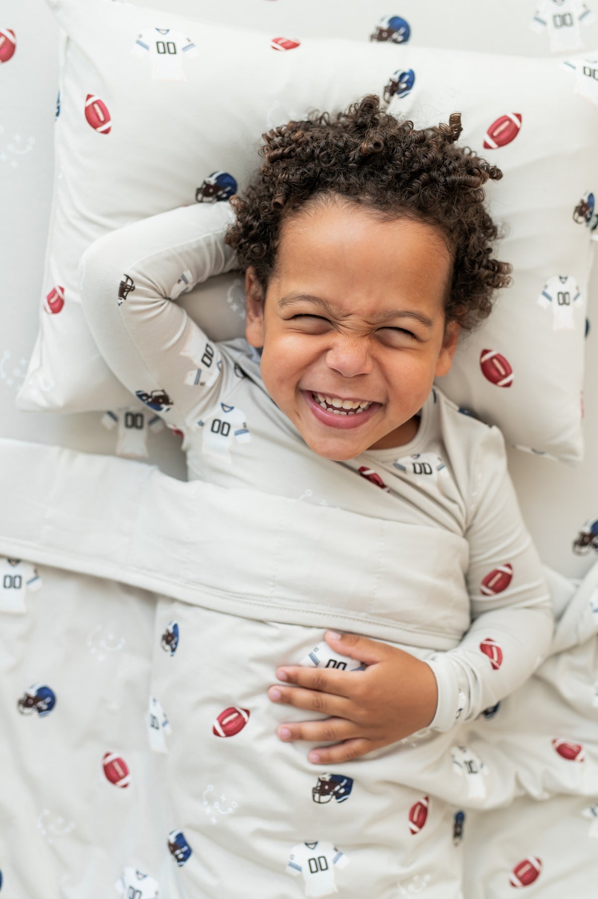 Boy lying on Toddler Pillowcase and sheet in Vintage Football print while wearing a similar print long sleeve pajama set covered with a similar print toddler blanket