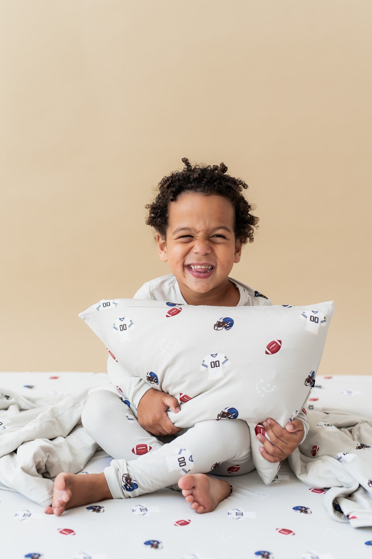 Boy holding Toddler Pillowcase in Vintage Football print sitting on similar print sheet while wearing a similar print long sleeve pajama set with a beige background