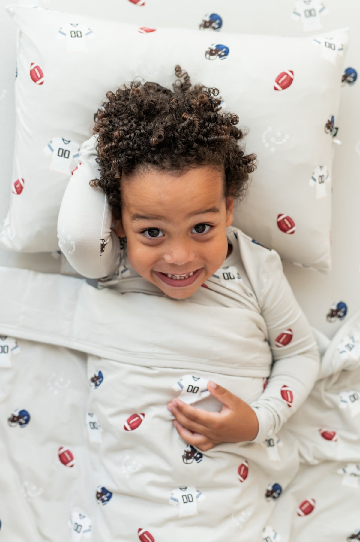 Boy lying on Toddler Pillowcase and sheet in Vintage Football print while wearing a similar print long sleeve pajama set covered with a similar print toddler blanket