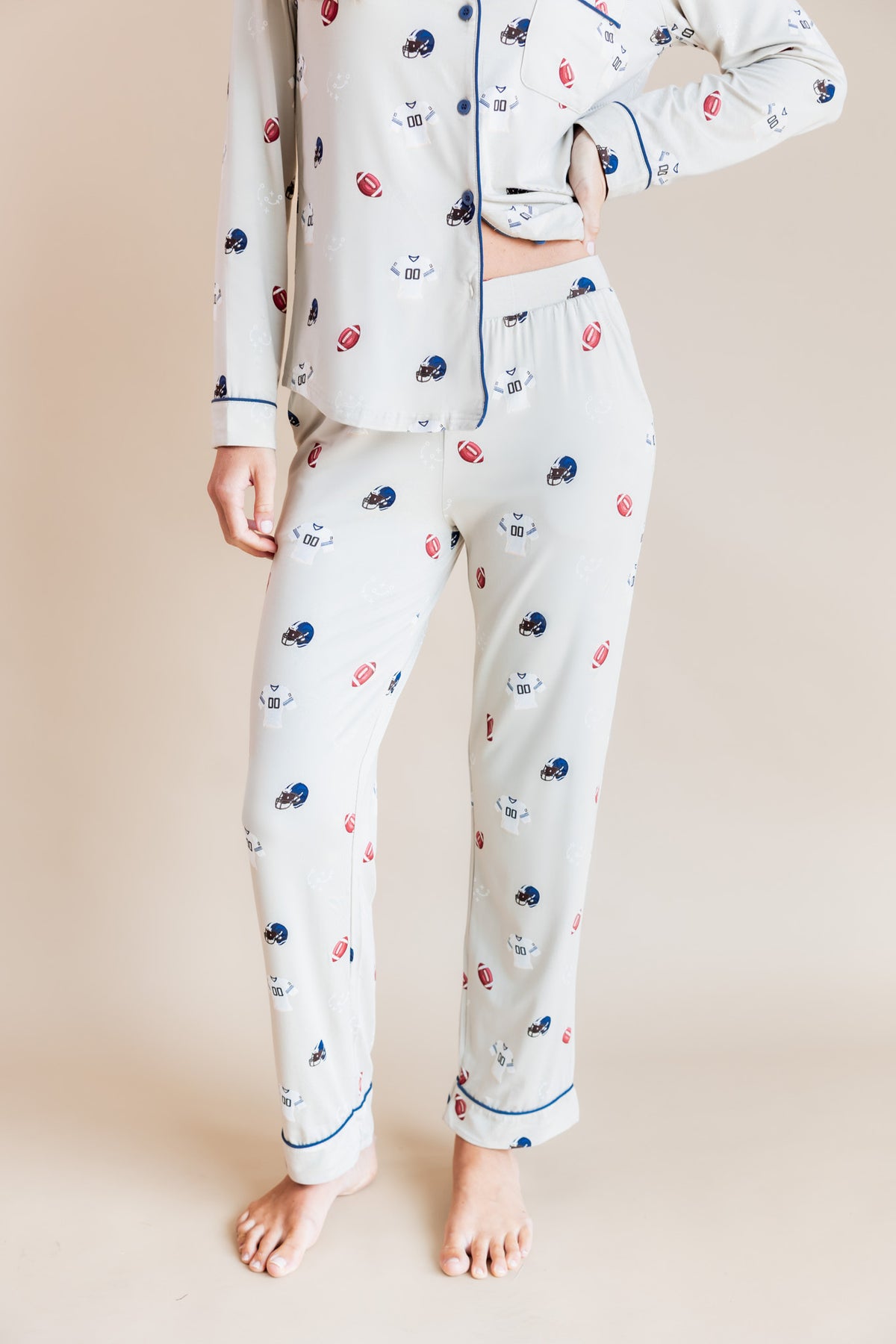 A model wearing a matching long-sleeve button-front pajama top and pants set in a warm off-white color, decorated with a small multicolor pattern of footballs, hearts, and stars, with blue piping details.