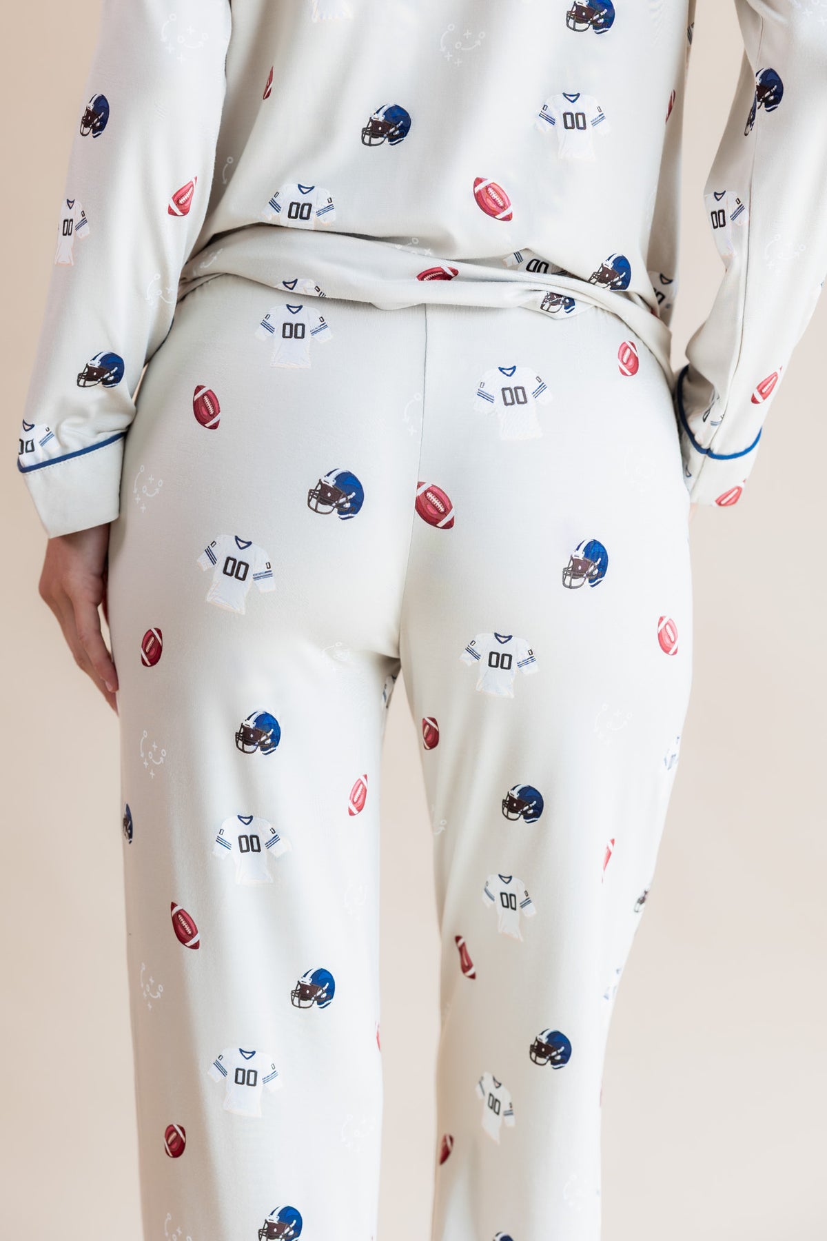 A person wearing a matching long-sleeve pajama top and pants set in an off-white color, both covered in a small repeating print of football helmets and footballs. The photo is cropped from the back, showing the waistband and upper legs against a plain light beige studio background.