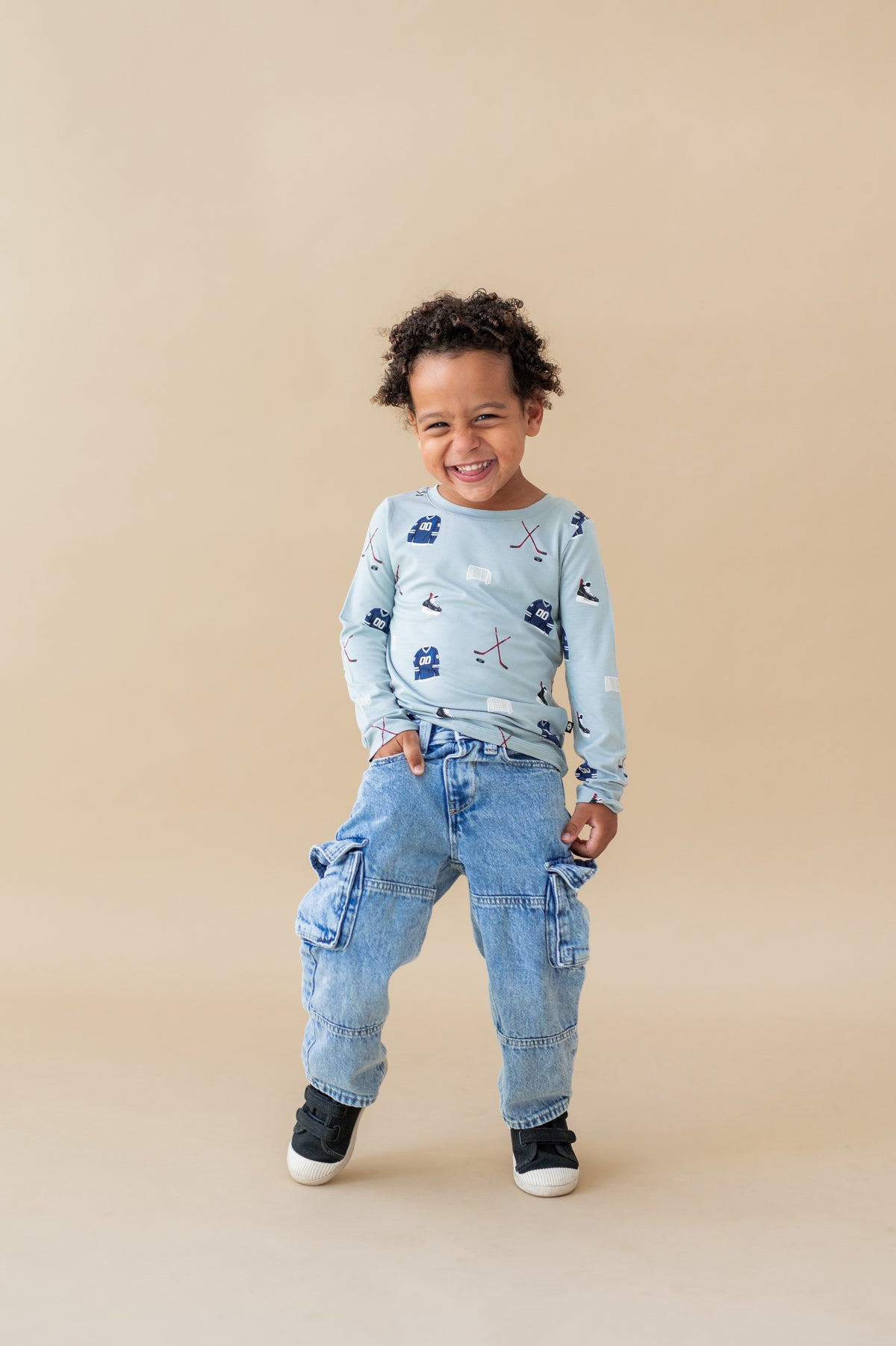 Boy wearing Long Sleeve Toddler Crew Neck Tee in Vintage Hockey print over blue jeans and black shoes on a beige background