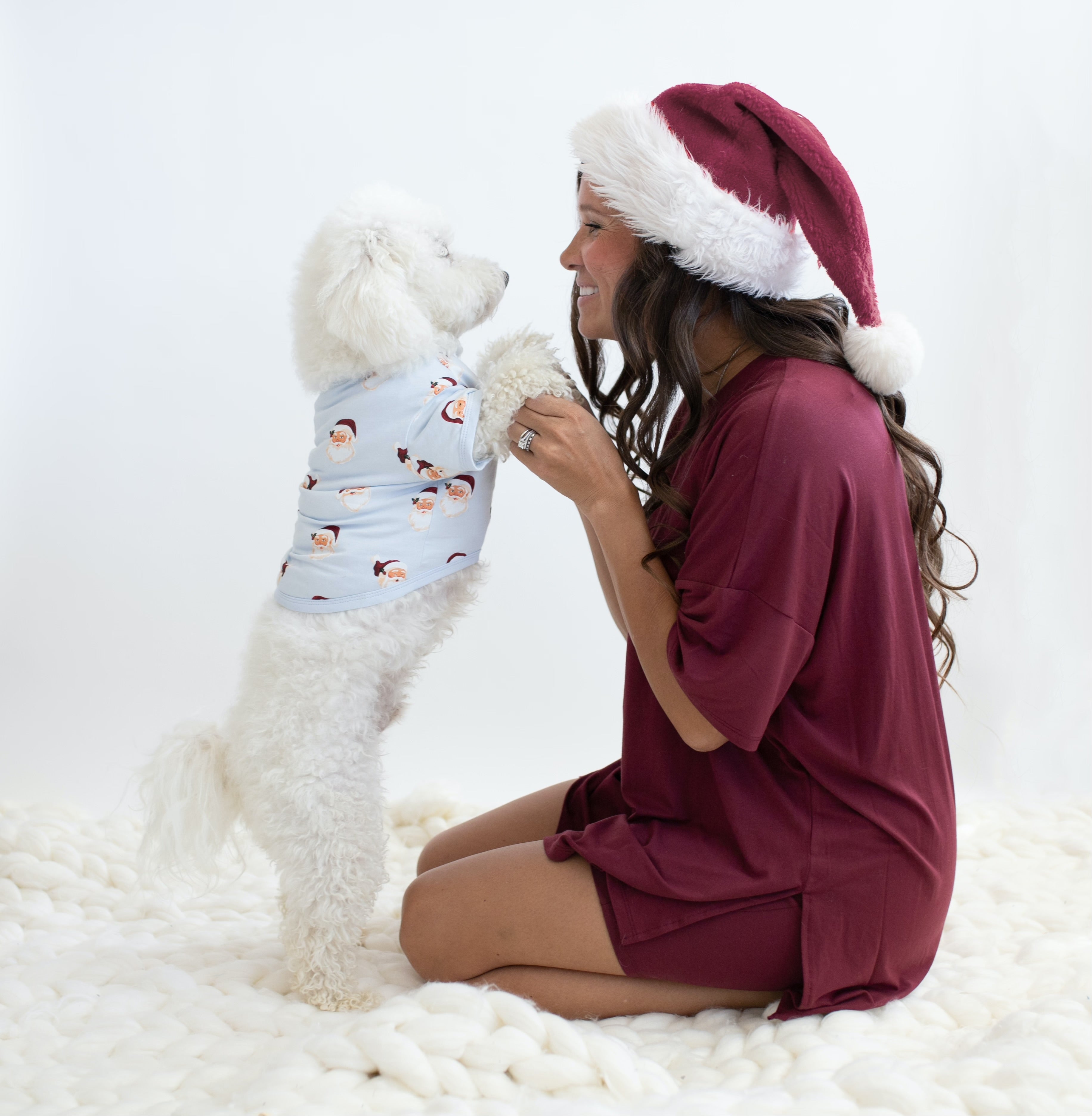 Female kneeling on the floor holding her dogs paws who is on their hind legs. Female model wearing the Women's Burgundy Biker set with a maroon colored santa hat while the dog is wearing the Dog Tee in Vintage Santa
