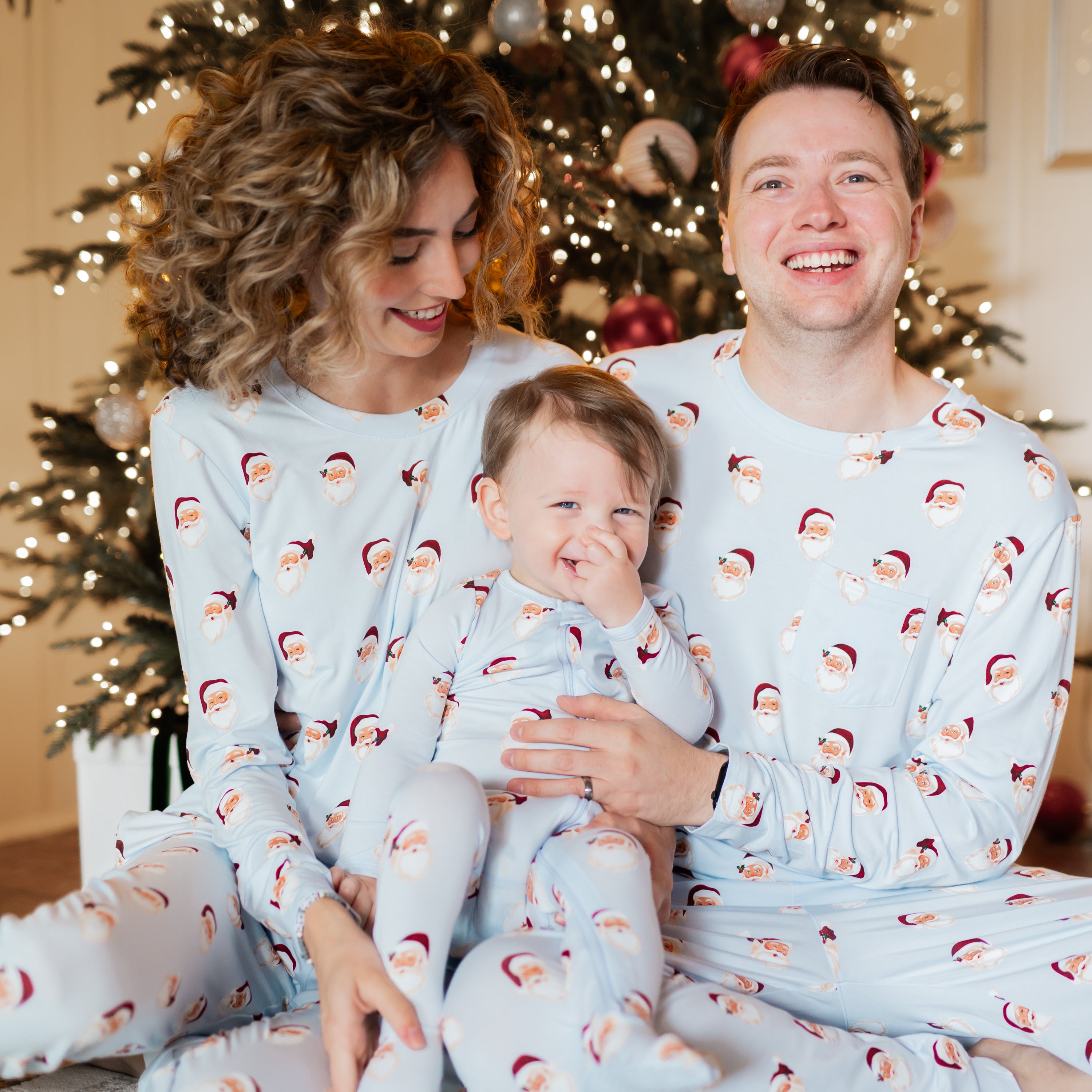 Family of three sitting on the floor in front of a decorated Christmas tree matching in the Vintage Santa print from the holiday collection