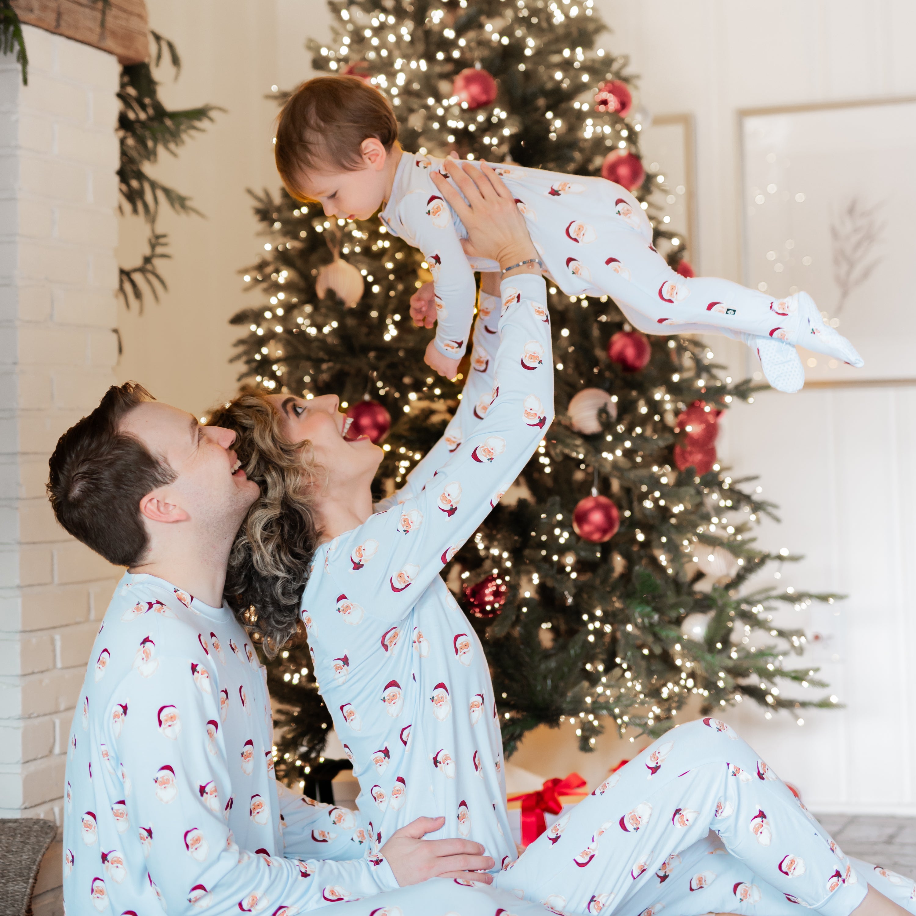 Mother and father sitting on the floor matching in Women's and Men's Jogger Pajama Set in Vintage Santa while wife is holding son up in the air who is matching in a zippered footie