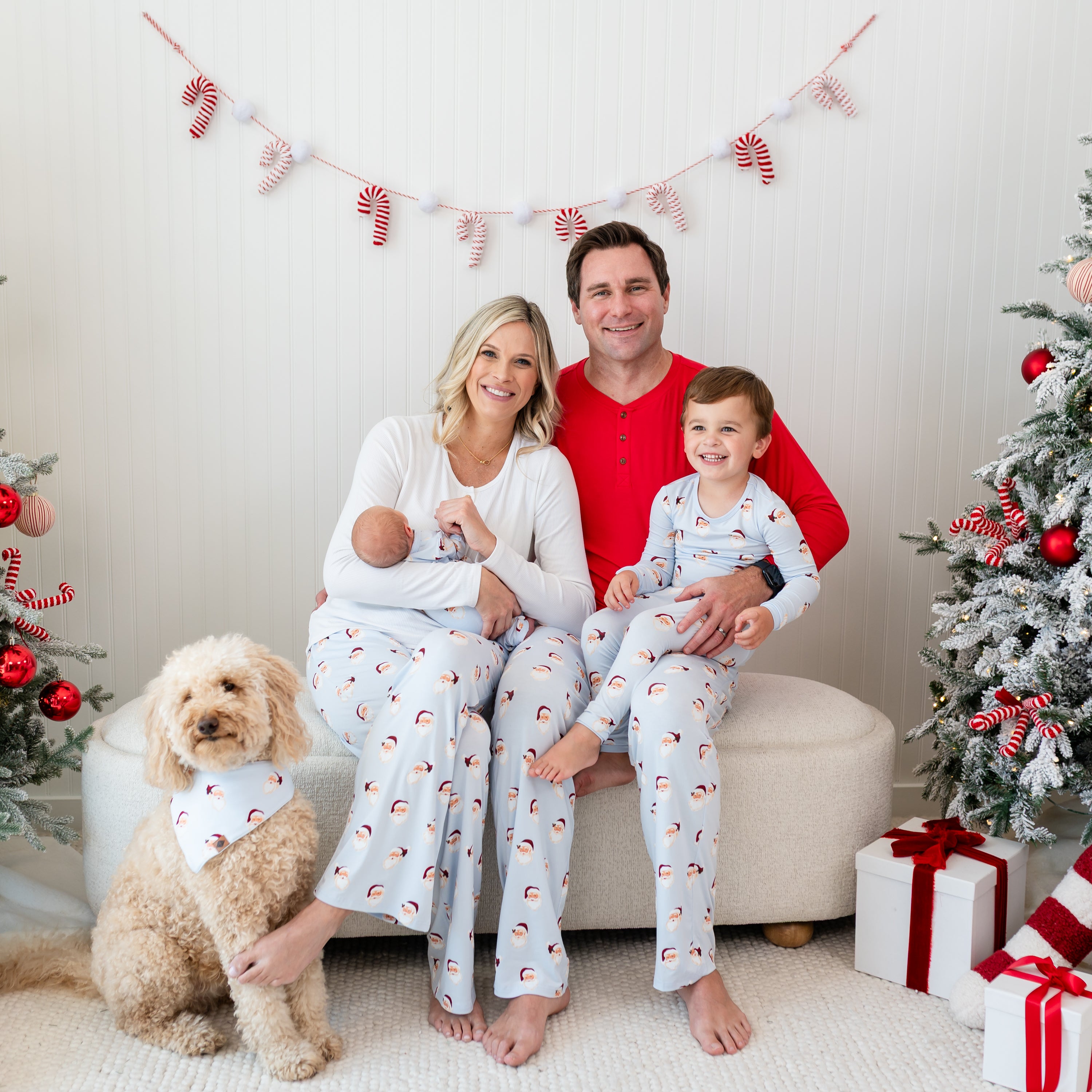Family of four sitting on a cream ottoman with a medium sized dog sitting in front of them all matching in various items in the Vintage Santa holiday print