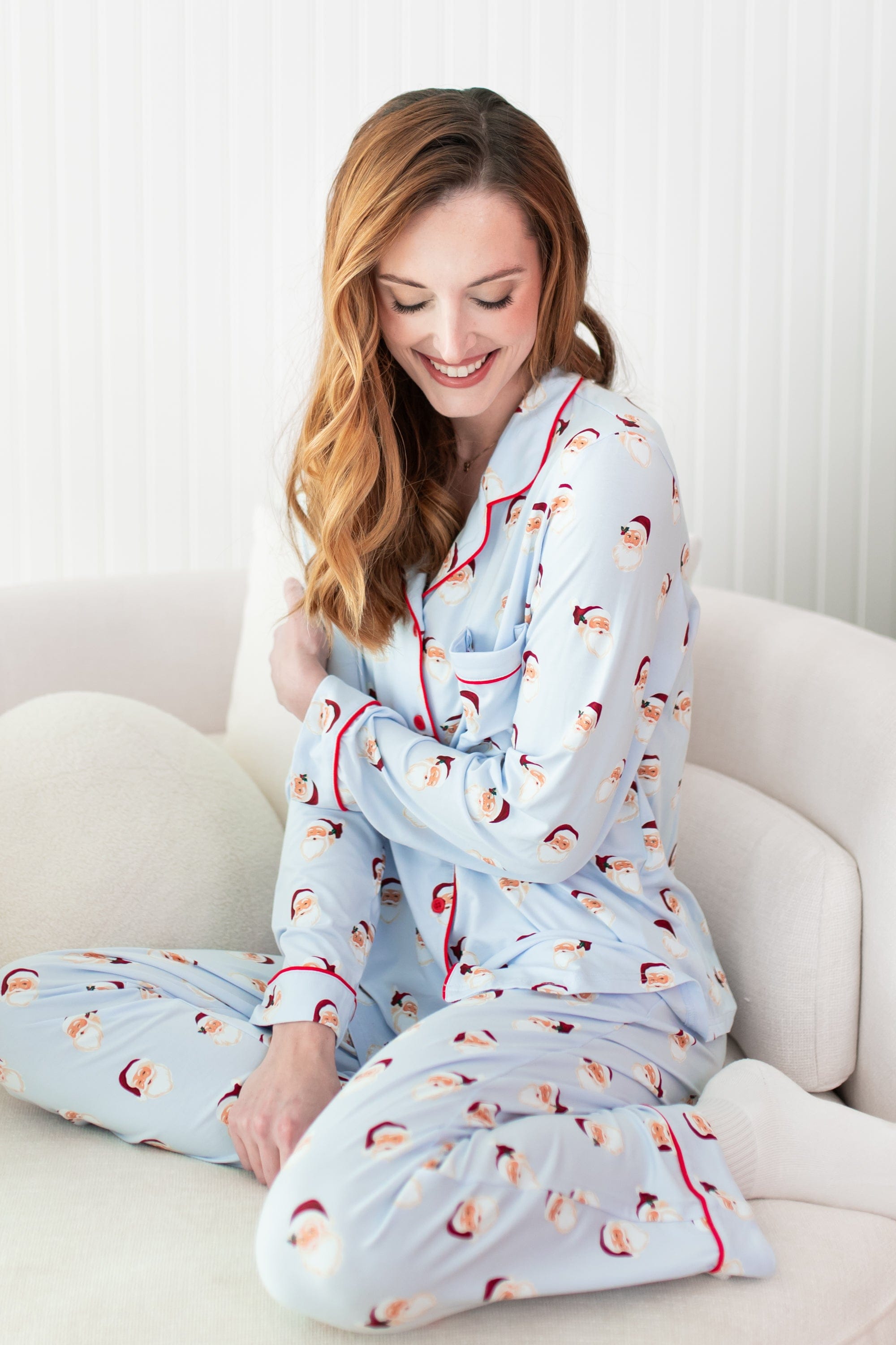Female model wearing the Long-Sleeved Women's Pajama Set in Vintage Santa sitting on a cream color sofa