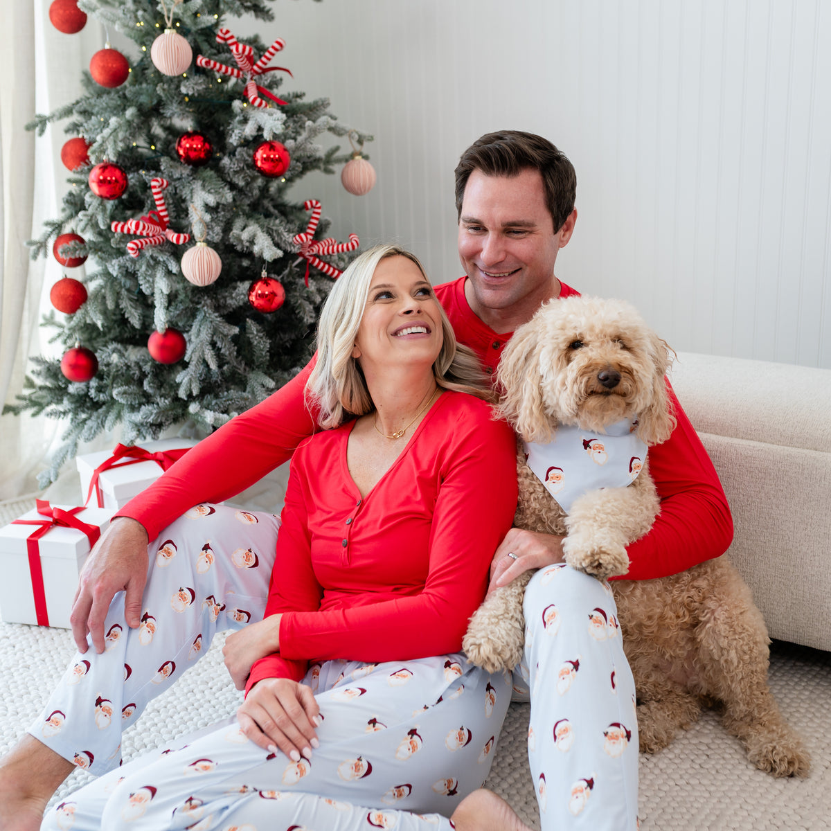 Husband and wife sitting on the floor wearing lounge pants in Lounge Pants in Vintage Santa with their dog beside them wearing a matching dog bandana