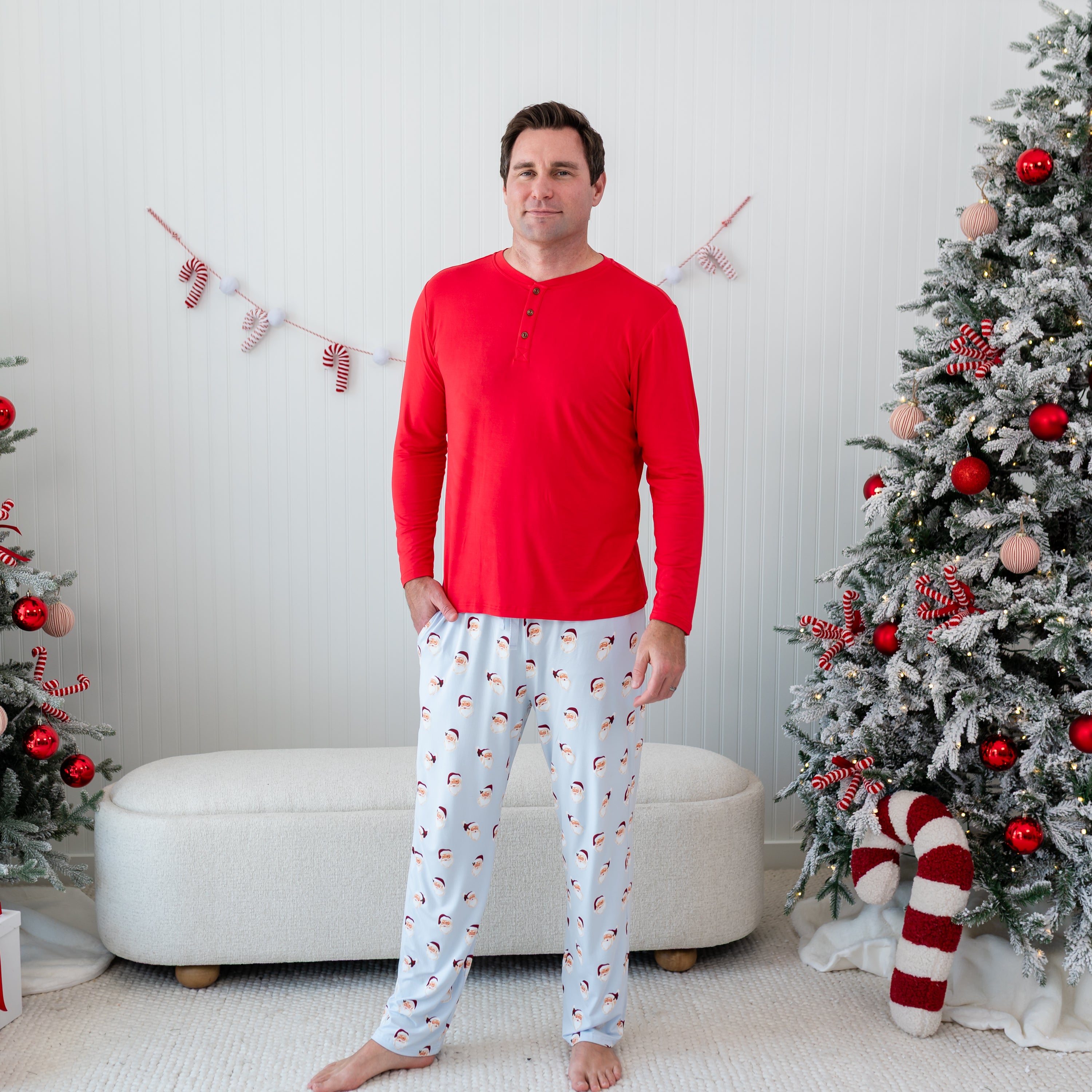 Male model standing in front of a cream colored ottoman and beside two decorated Christmas trees wearing the Men's Lounge Pants in Vintage Santa and a Men's Henley top in Cardinal