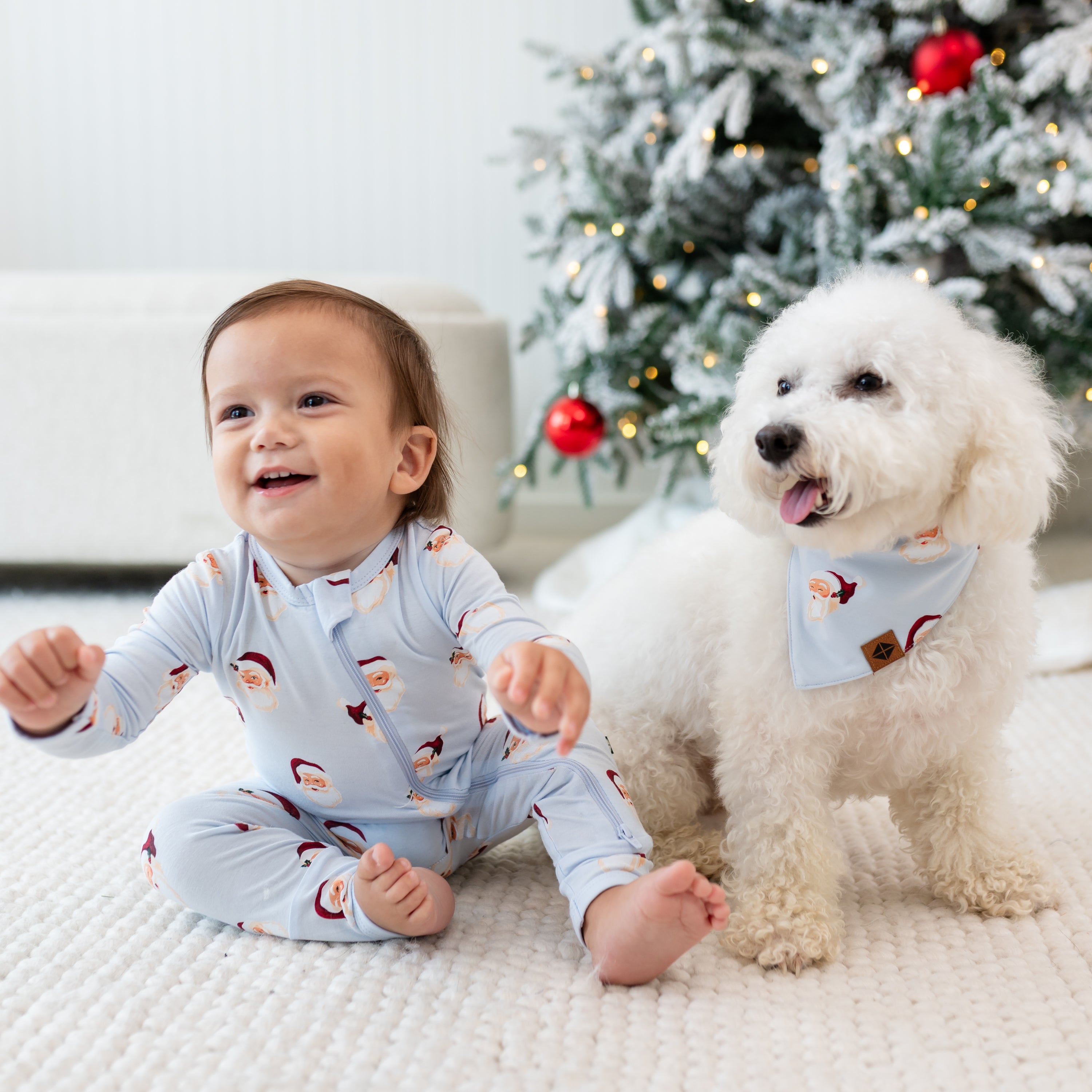 Small white dog wearing the Dog Bandana in Vintage Santa sitting beside a young toddler who is wearing a matching zippered romper