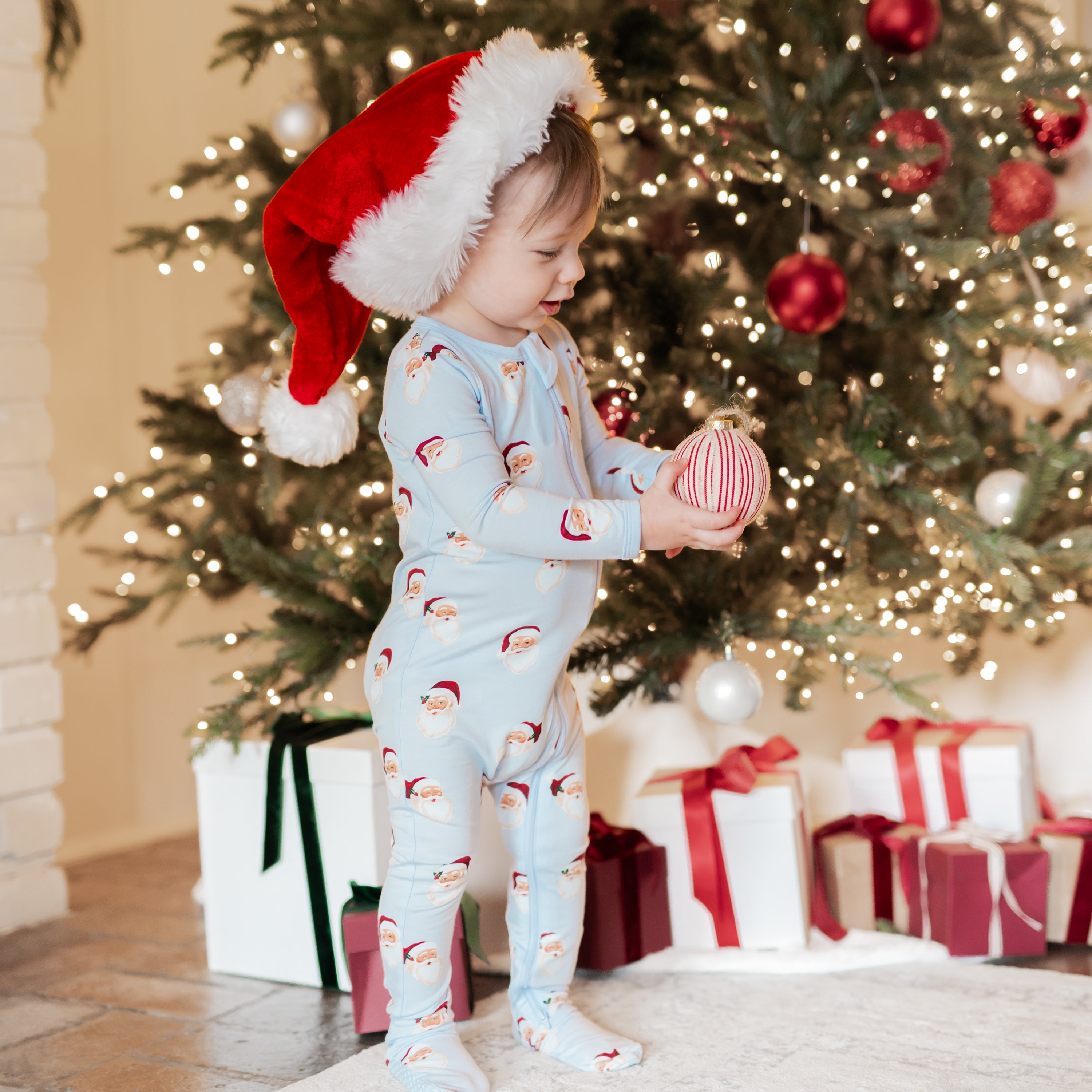 Young boy standing in front of a Christmas tree holding a christmas ornament in his hand wearing the Zippered Footie in Vintage Santa with a santa hat