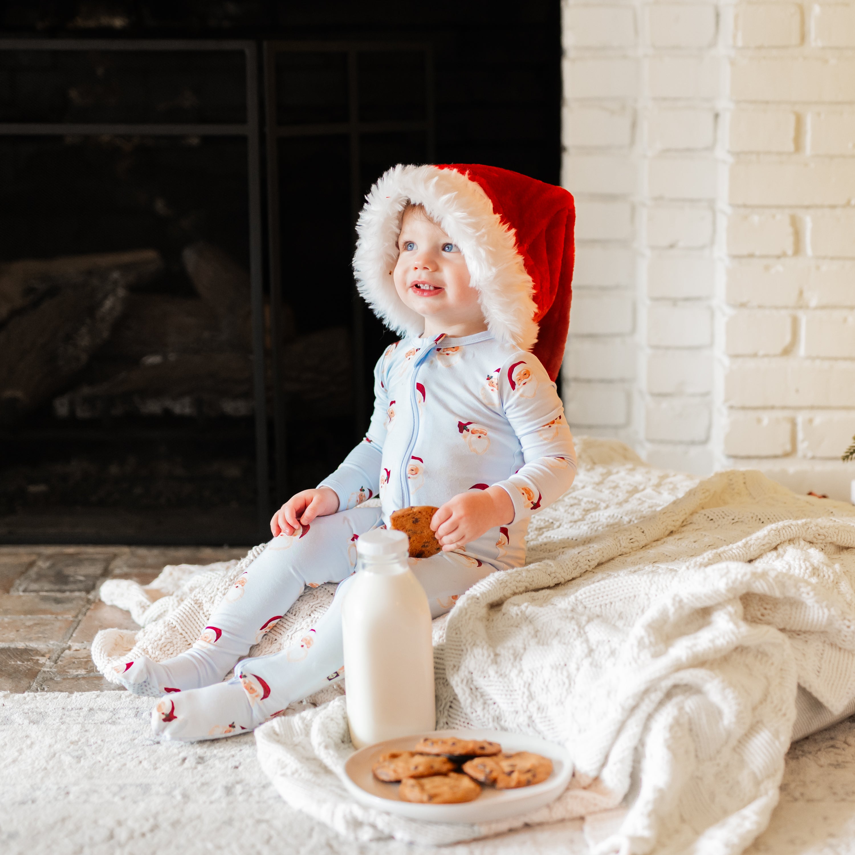 Young boy sitting on the floor with a cookie in his hand and a glass milk jug and plate of cookies in front of him wearing the Zippered Footie in Vintage Santa with a santa hat