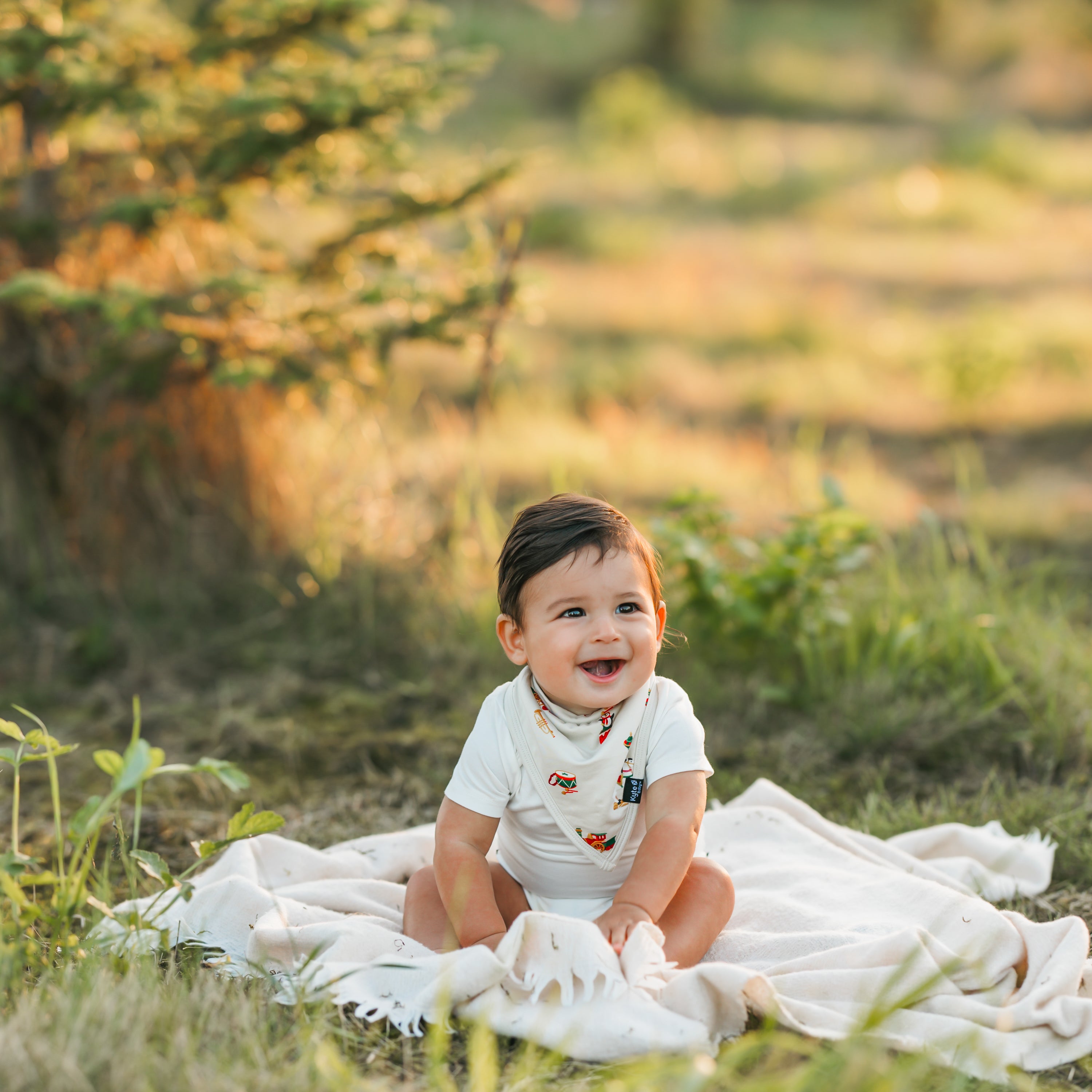 Smiling toddler sitting on a white blanket in the grass wearing the Bib in Vintage Toys