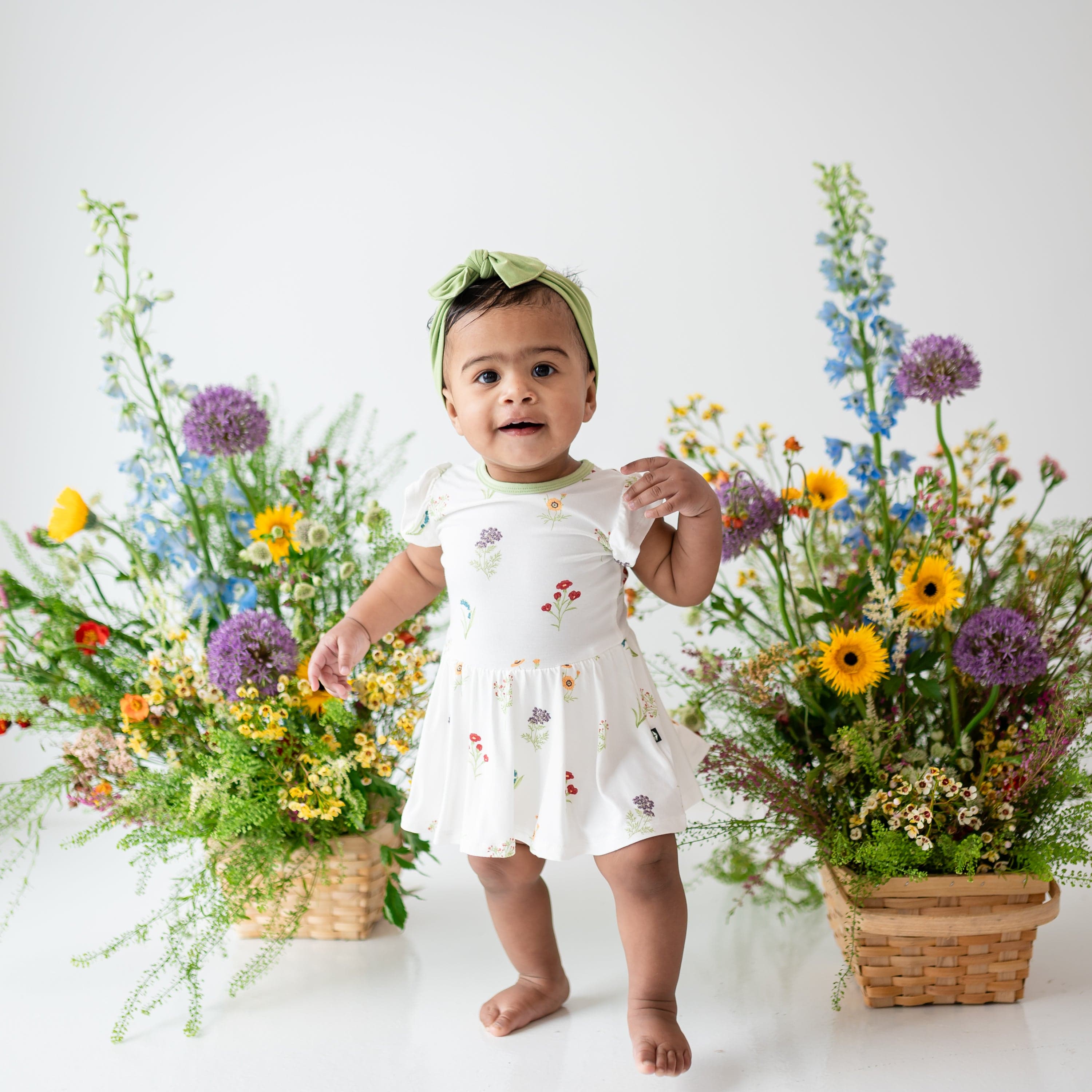 Standing young toddler modeling Twirl Bodysuit Dress in Wildflower next to flowers