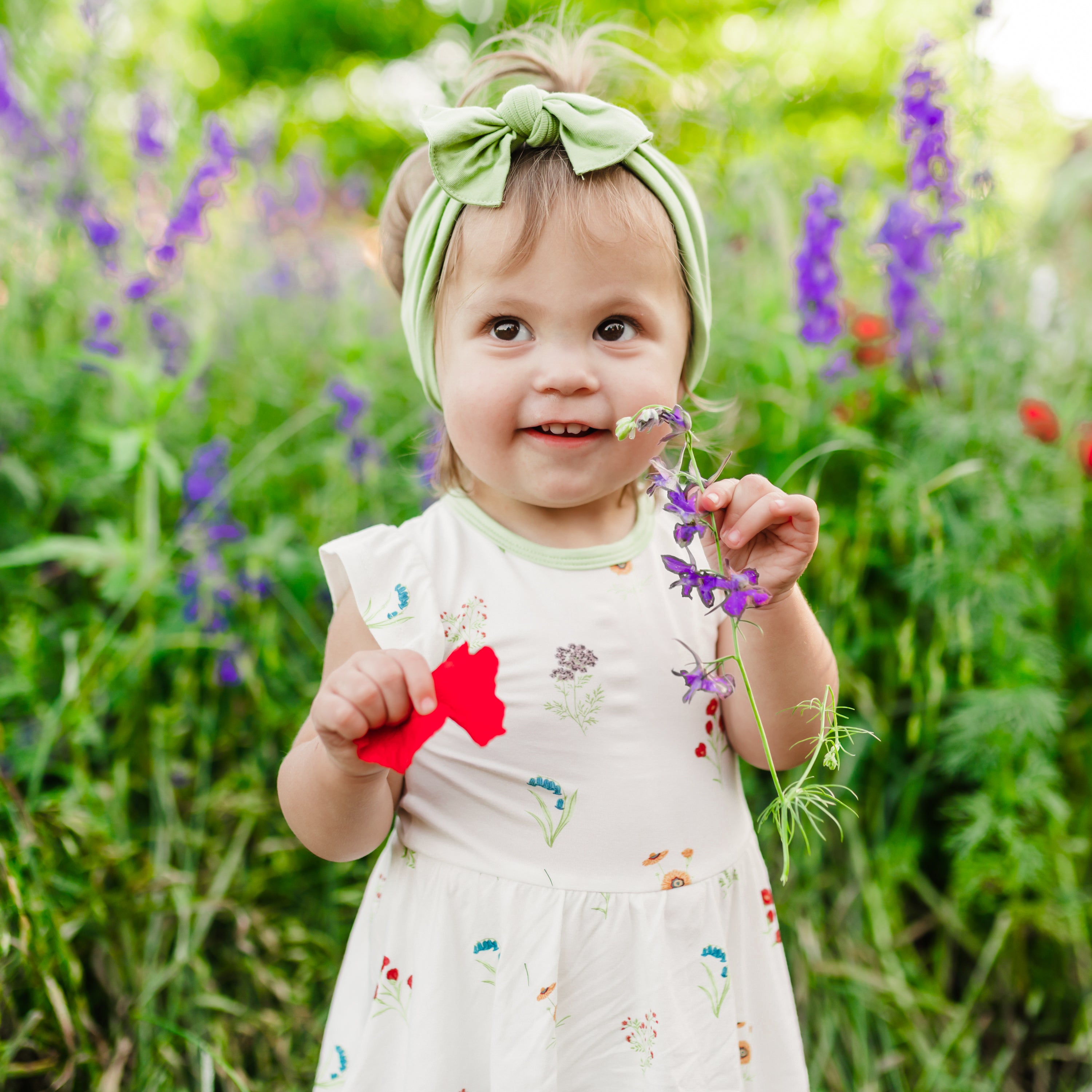 Toddler wearing Twirl Bodysuit Dress in Wildflower in a field of flowers smelling flower