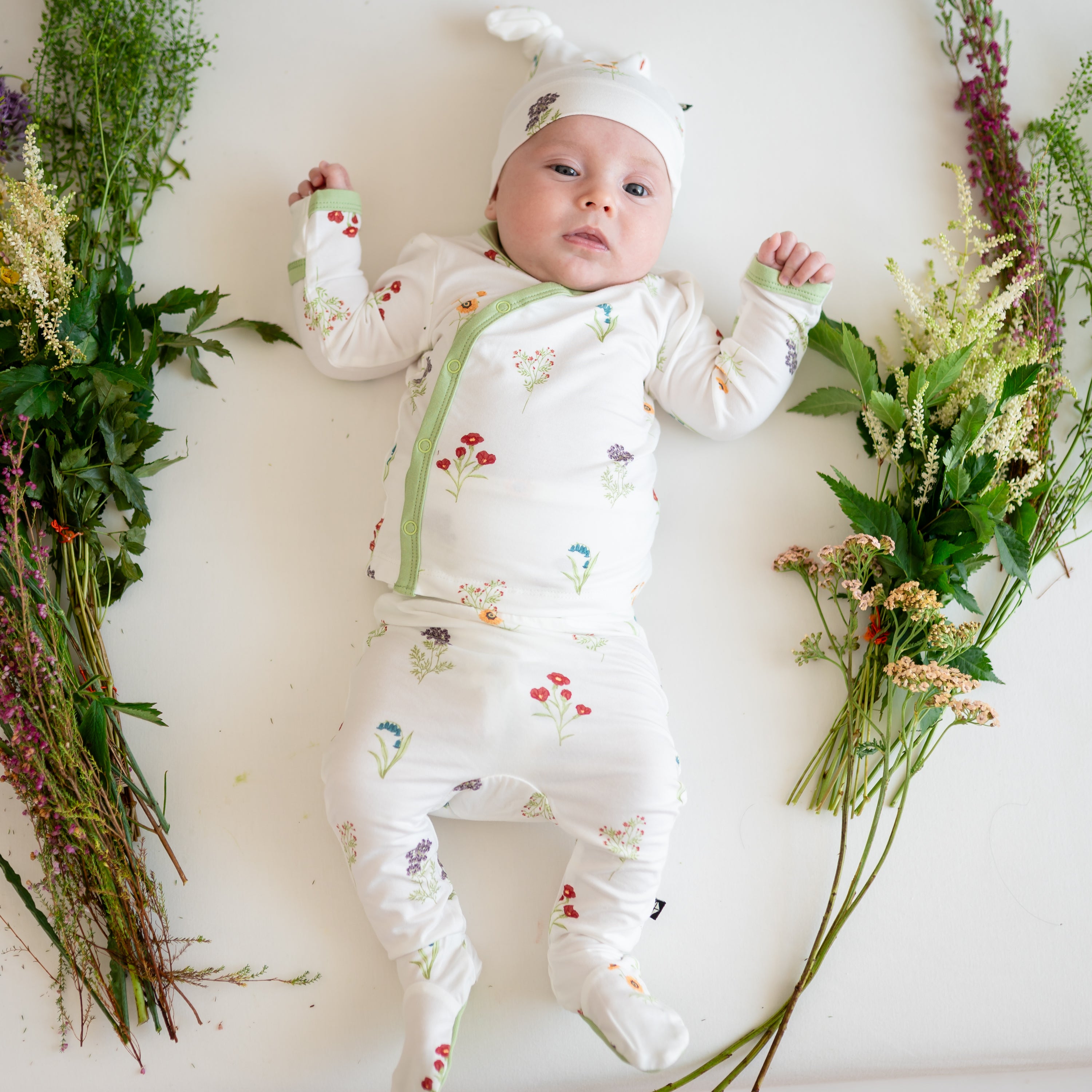 Baby wearing Take Me Home Set with Cap in Wildflower surrounded by flowers