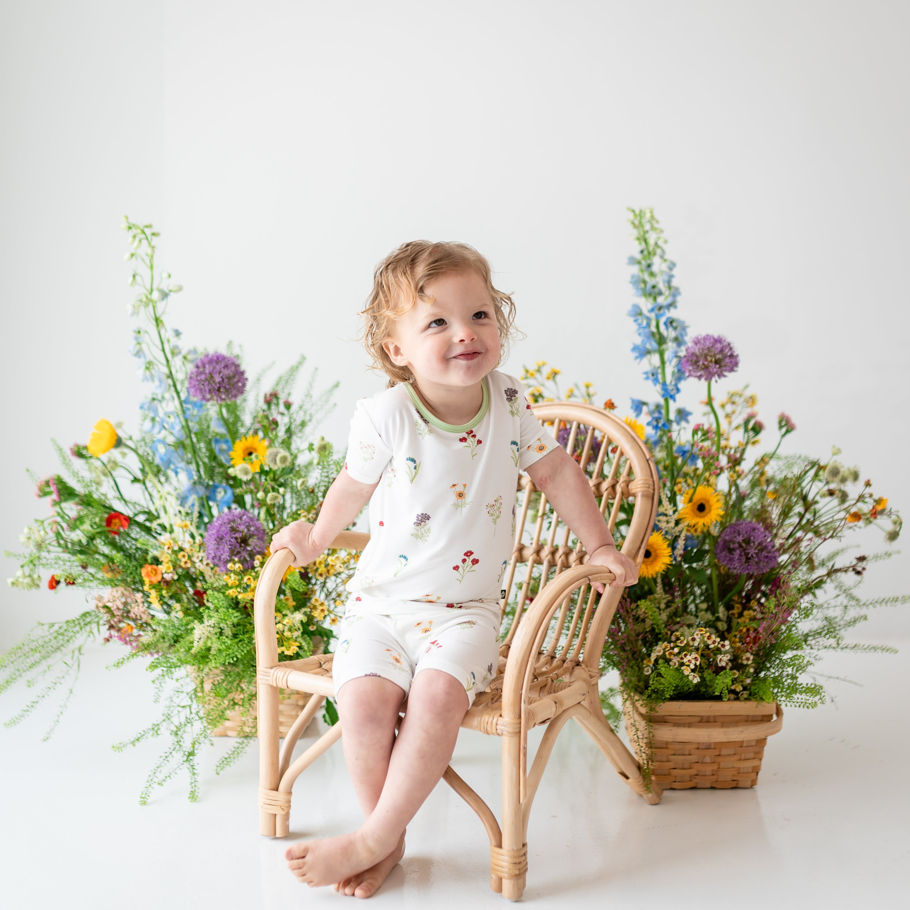Toddler wearing Short Sleeve Pajamas in Wildflower with a soft smile on a wooden chair with flower in background