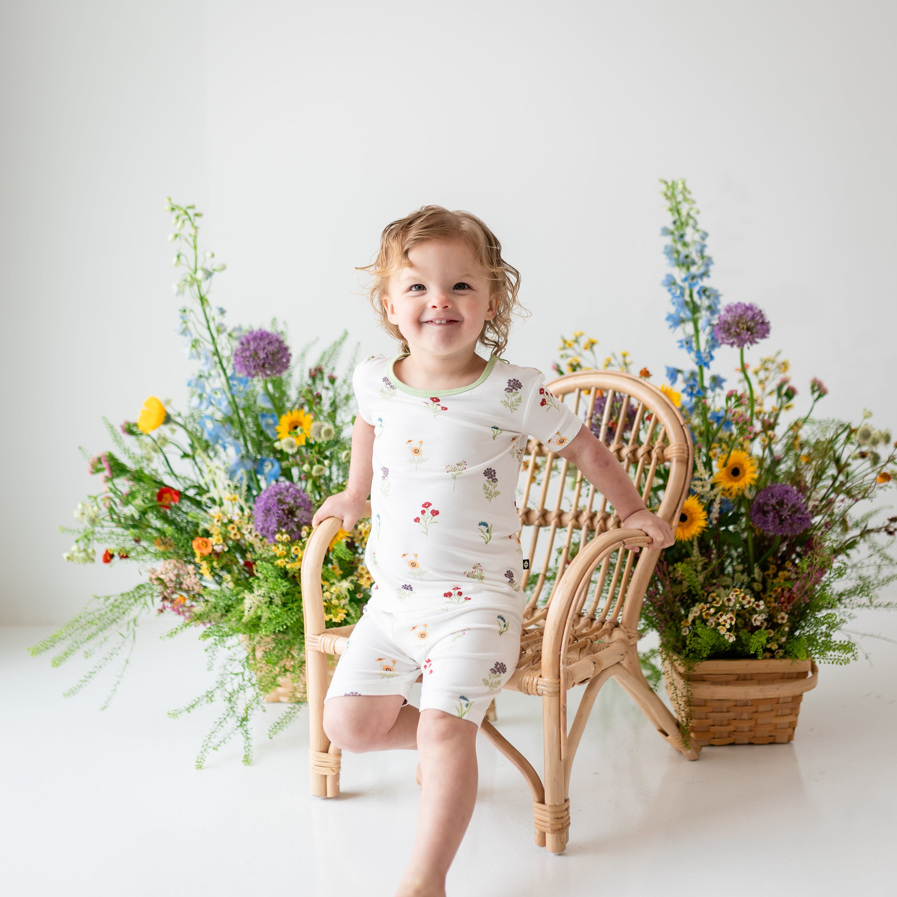 Toddler modeling Short Sleeve Pajamas in Wildflower on a wooden chair with flower in background