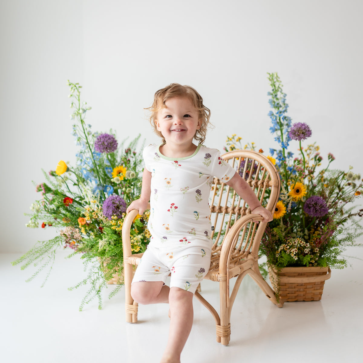 Toddler modeling Short Sleeve Pajamas in Wildflower on a wooden chair with flower in background