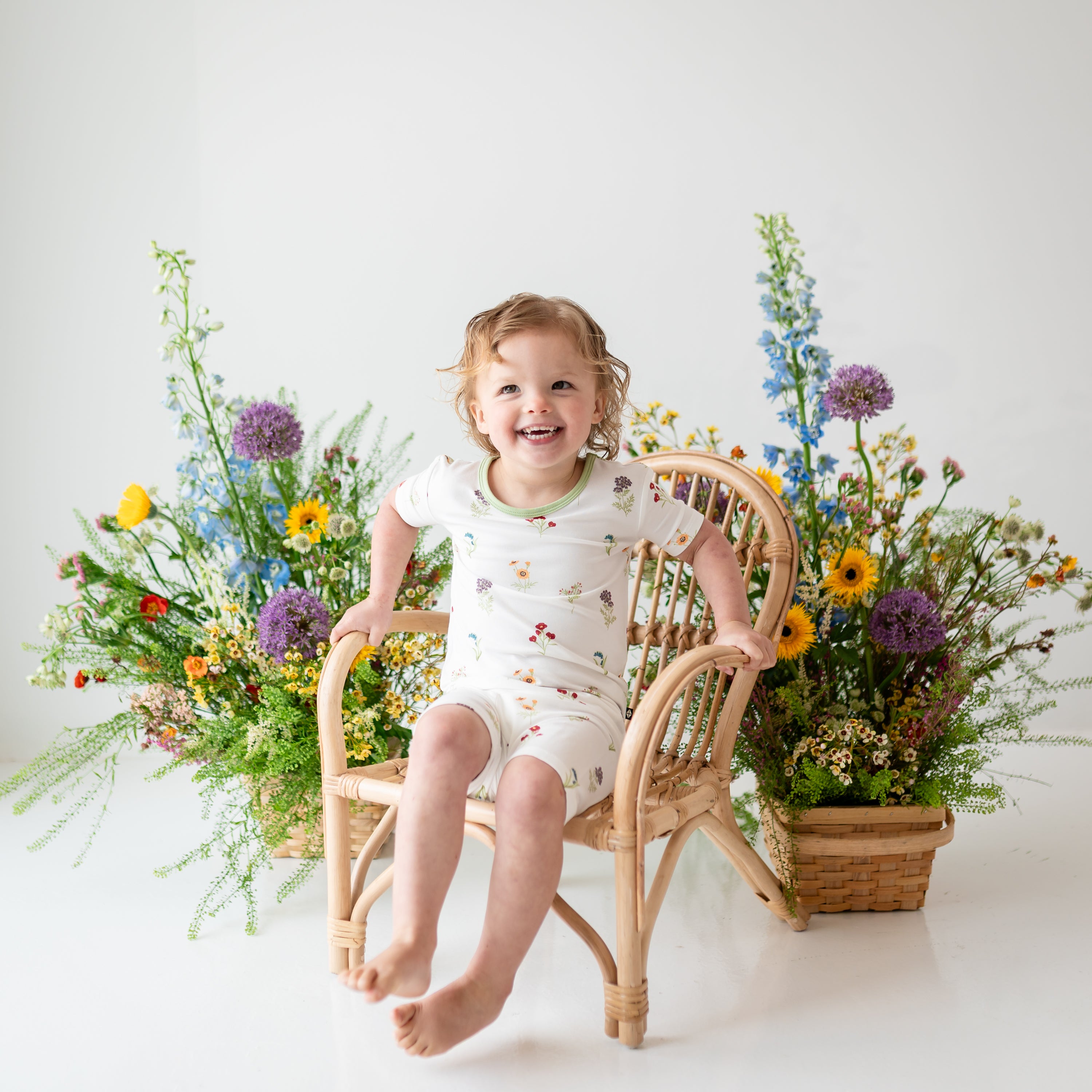 Toddler laughing wearing Short Sleeve Pajamas in Wildflower on a wooden chair with flower in background