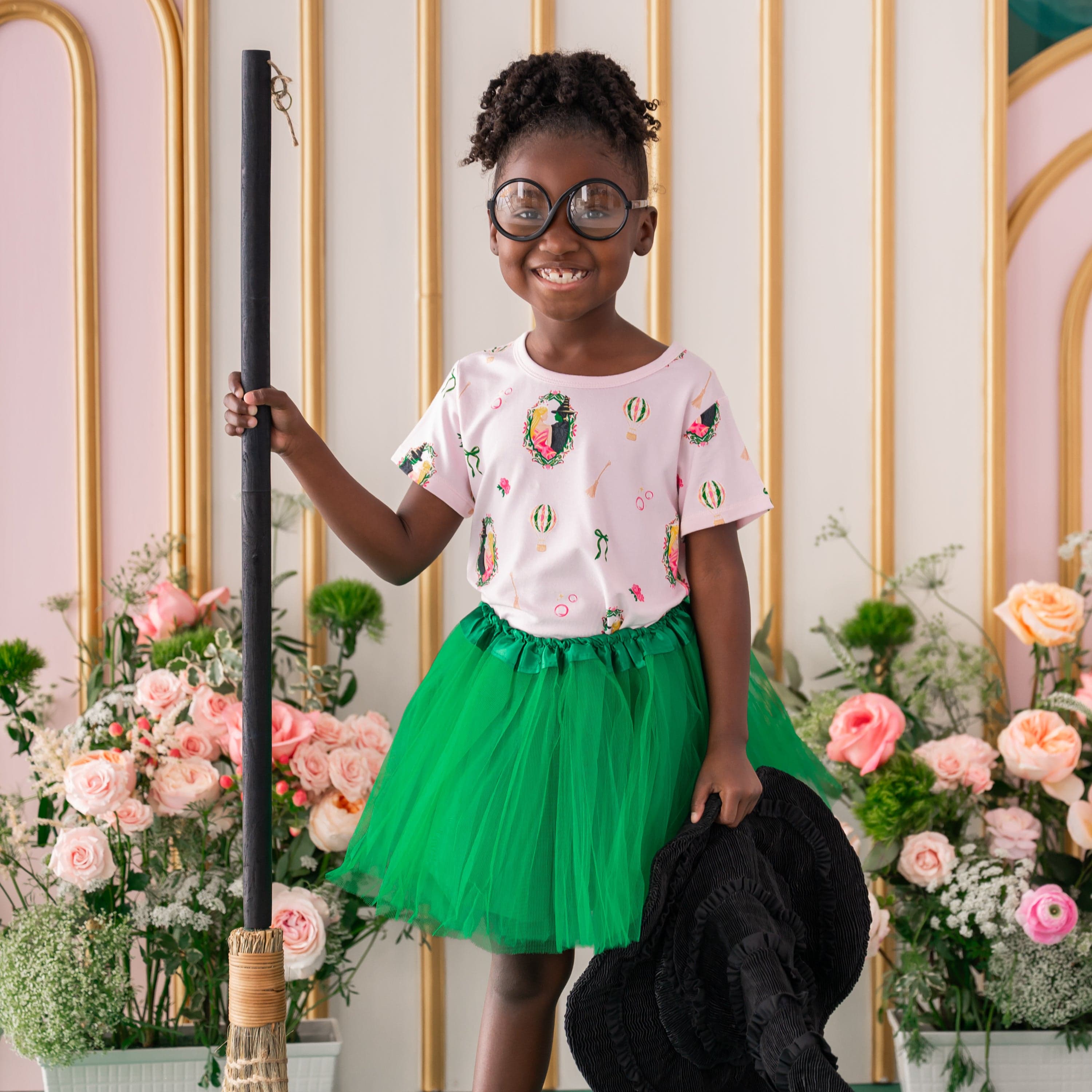 Smiling girl standing holding a broom and black witches hat wearing circle prop glasses and Toddler Basic Tee in Wicked with a green tulle skirt