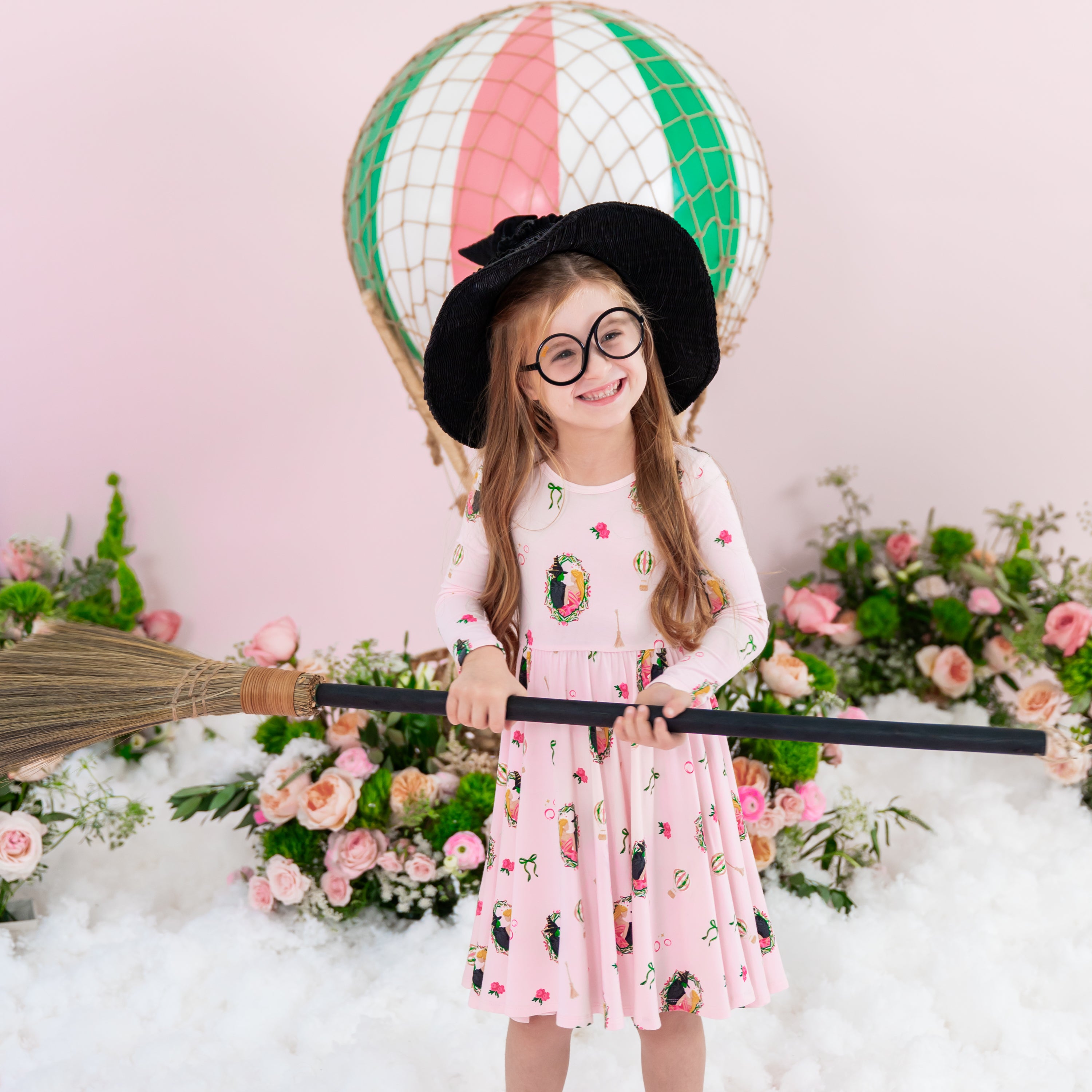 Young smiling girl holding a broom wearing a black witches hat wearing the soft and breathable Long Sleeve Twirl Dress in Wicked from Kyte Baby
