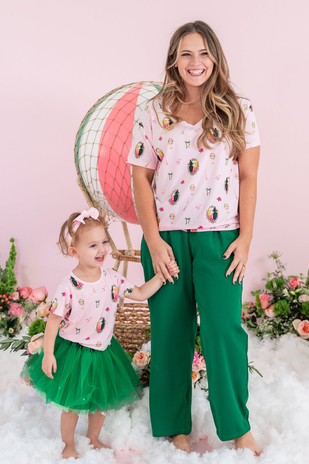 Mother wearing the Women's Relaxed Fit V-Neck in Wicked and green pants holding her daughters hand who is matching in a basic tee and green tulle skirt standing in front of a prop hot air balloon and flowers