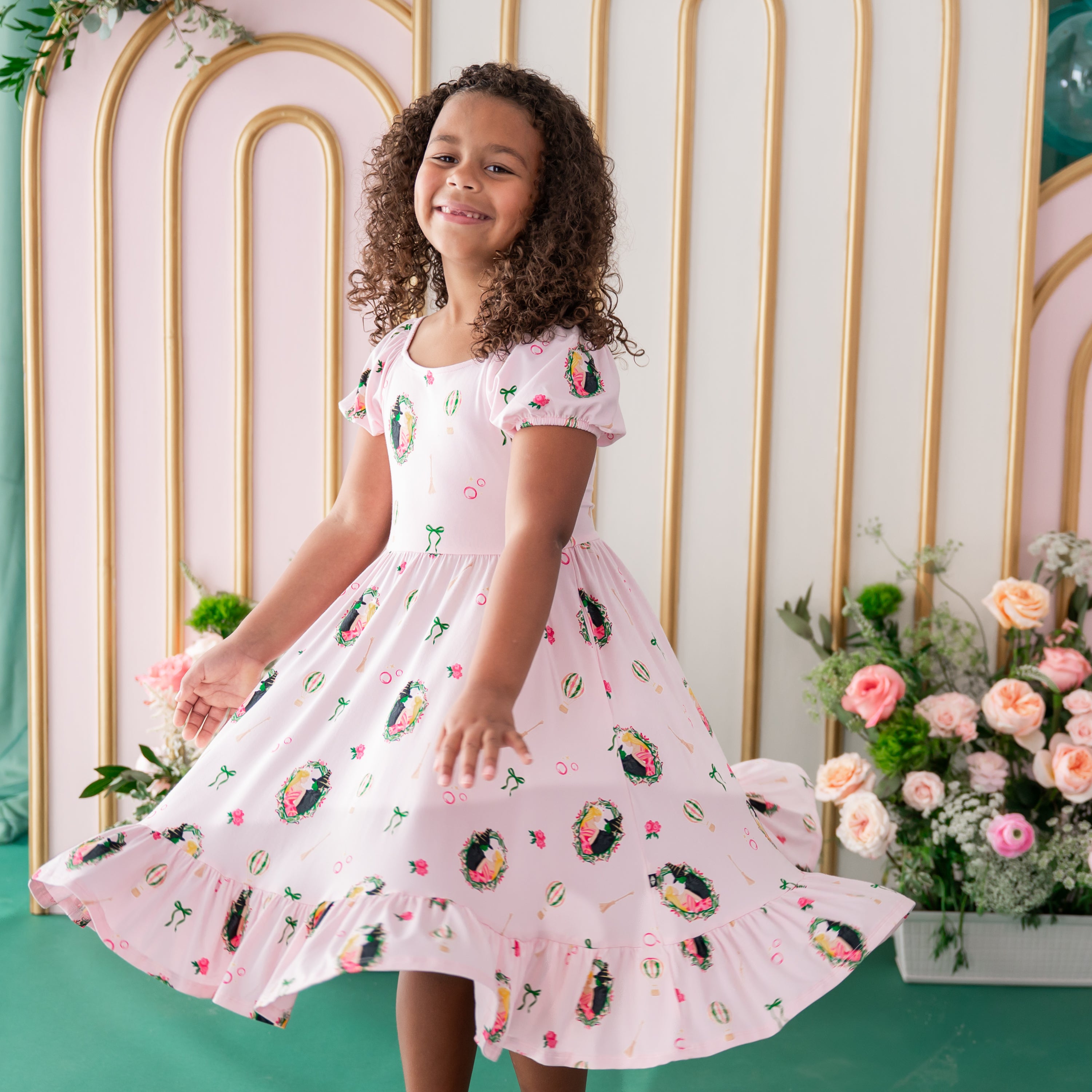 Smiling girl twirling in the soft and breathable Puff Sleeve Twirl Dress in Wicked made by Kyte Baby standing in front of a floral backdrop