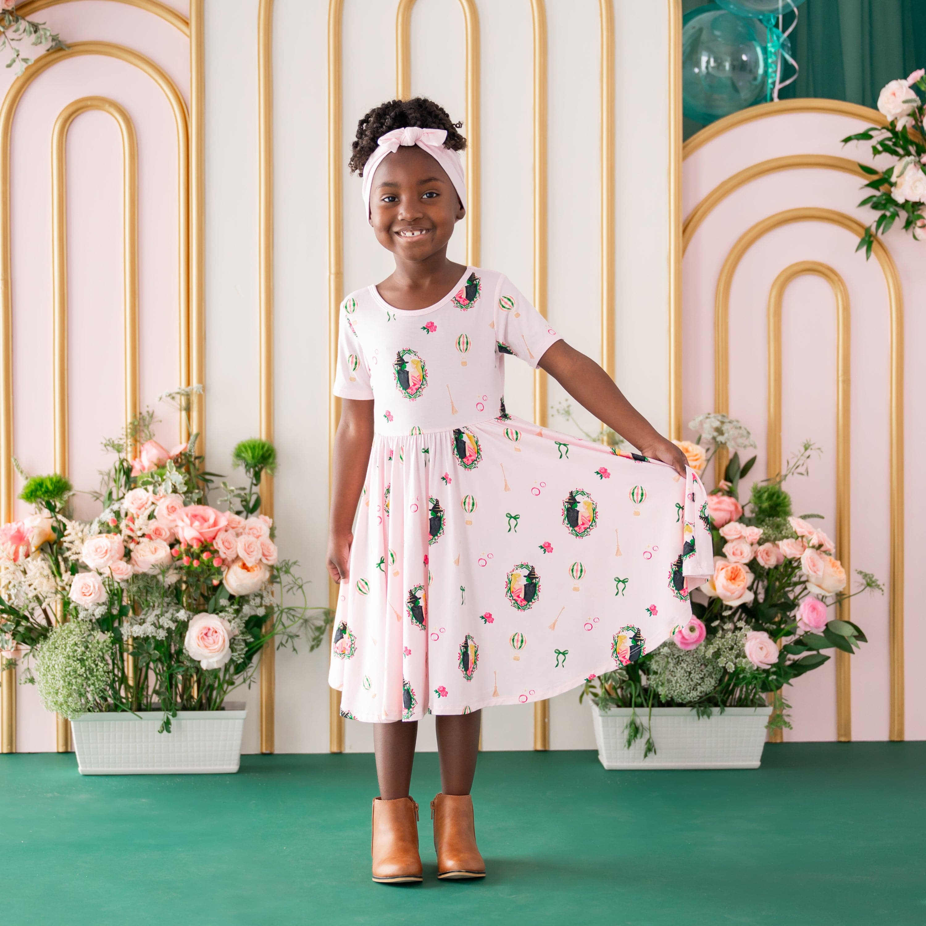Young girl standing in front of a floral backdrop holding the hem of the Twirl Dress in Wicked