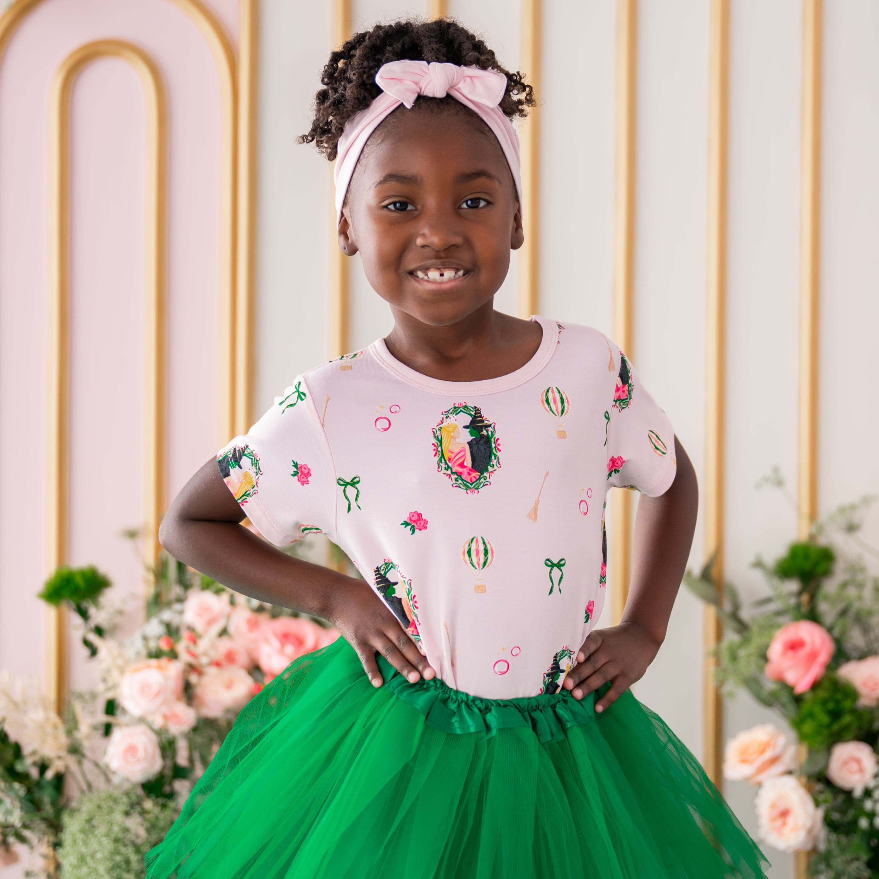 Smiling model wearing the Toddler Basic Tee in Wicked with a green tulle skirt standing in front of a floral backdrop