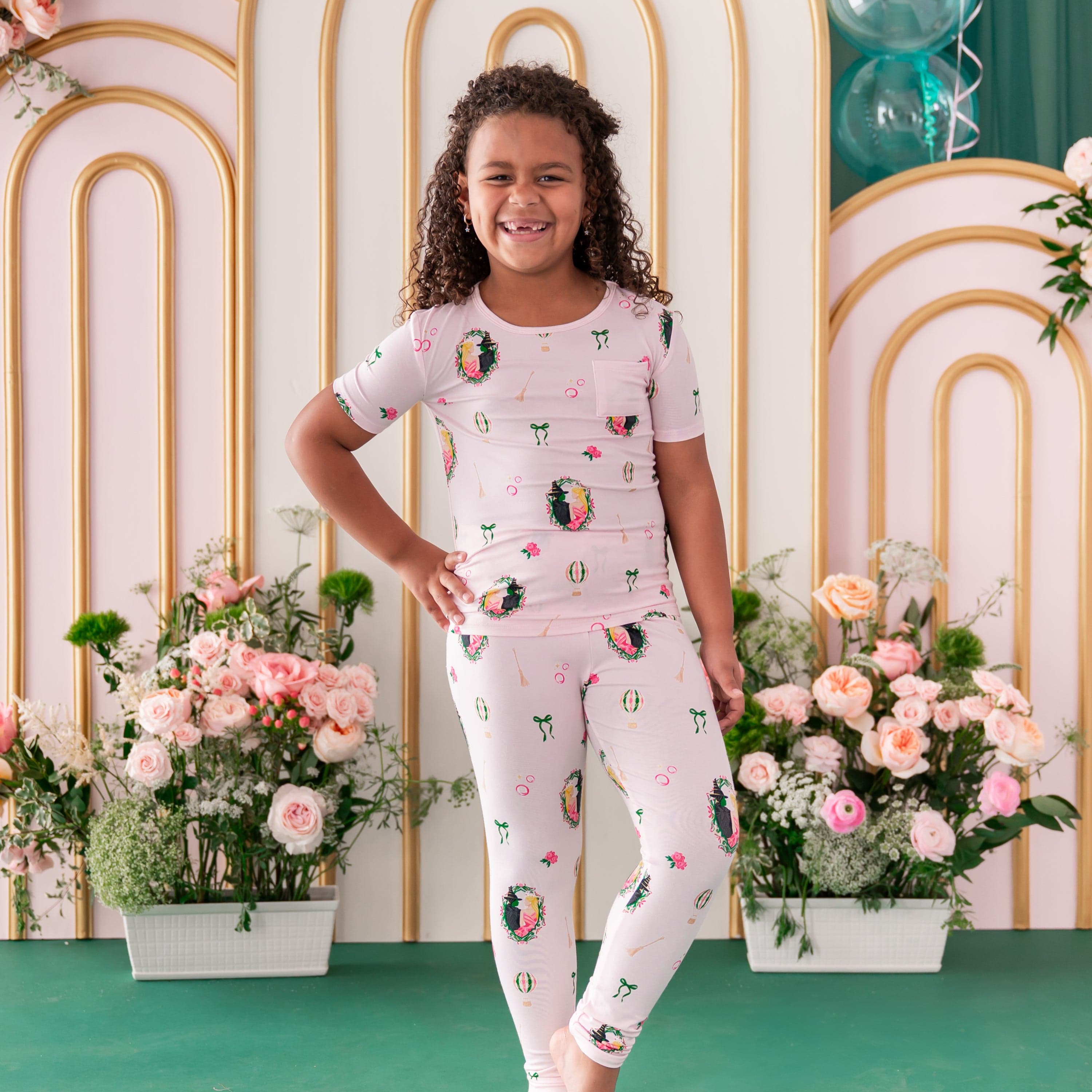 Young smiling girl standing with her hand on her hip wearing the soft and breathable Short Sleeve with Pants Pajamas in Wicked from Kyte Baby standing in front of a floral backdrop