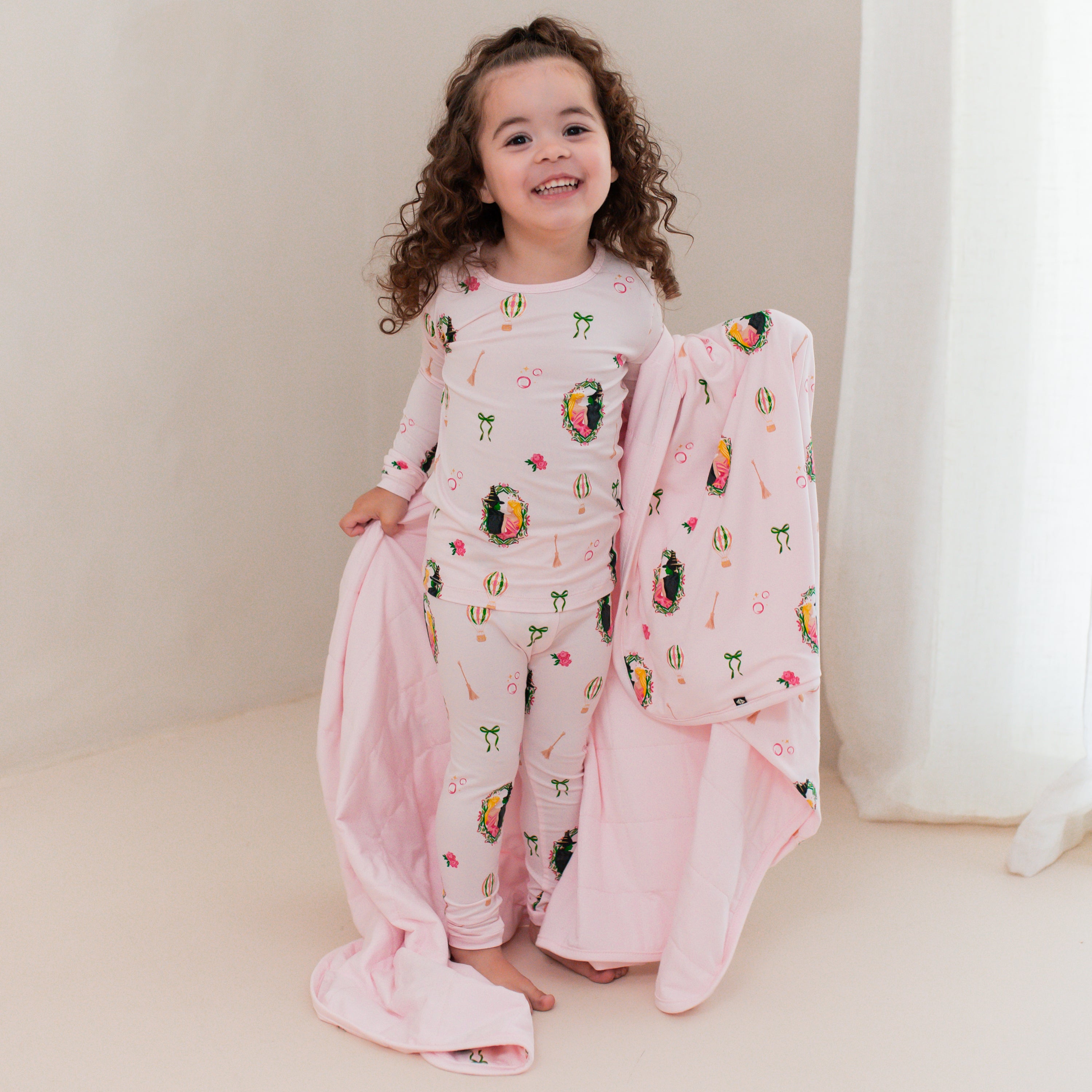 Young girl standing in front of a beige wall wearing the Long Sleeve Pajamas in Wicked holding a matching toddler blanket