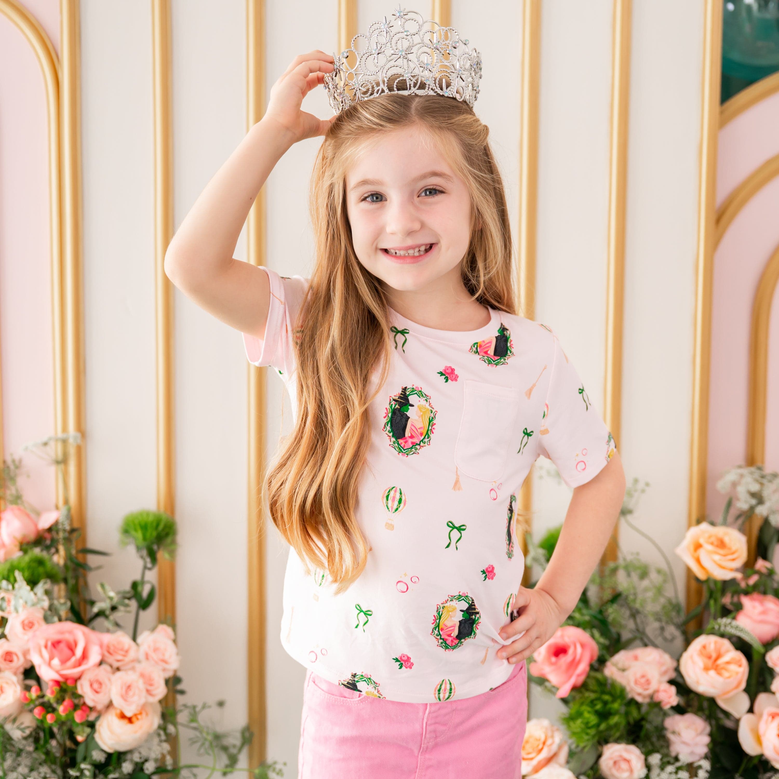 Smiling girl wearing the Toddler Crew Neck Tee in Wicked and a tiara standing in front of a floral backdrop