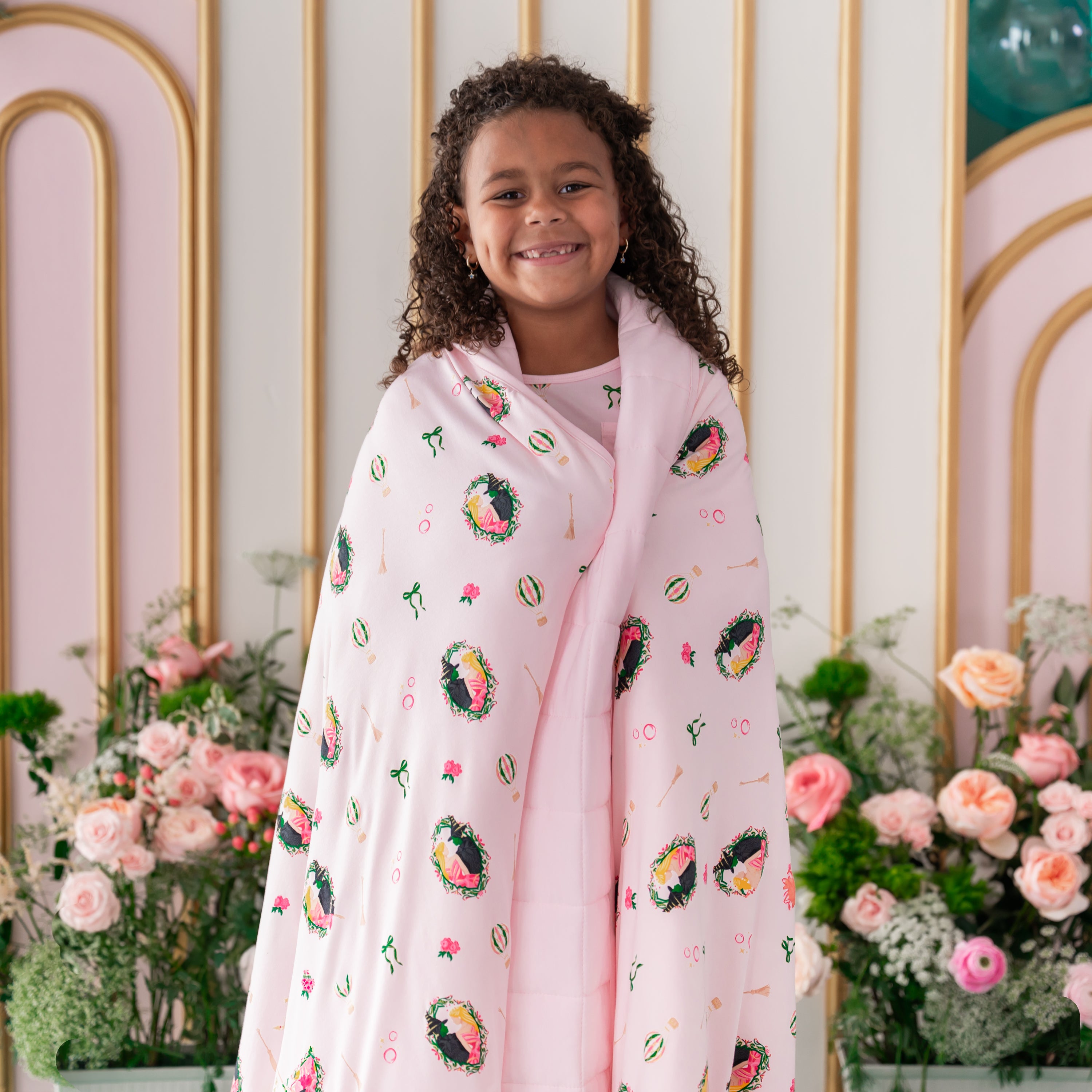 Smiling girl standing in front of a floral backdrop with the Youth Blanket in Wicked 2.5 wrapped around her shoulders