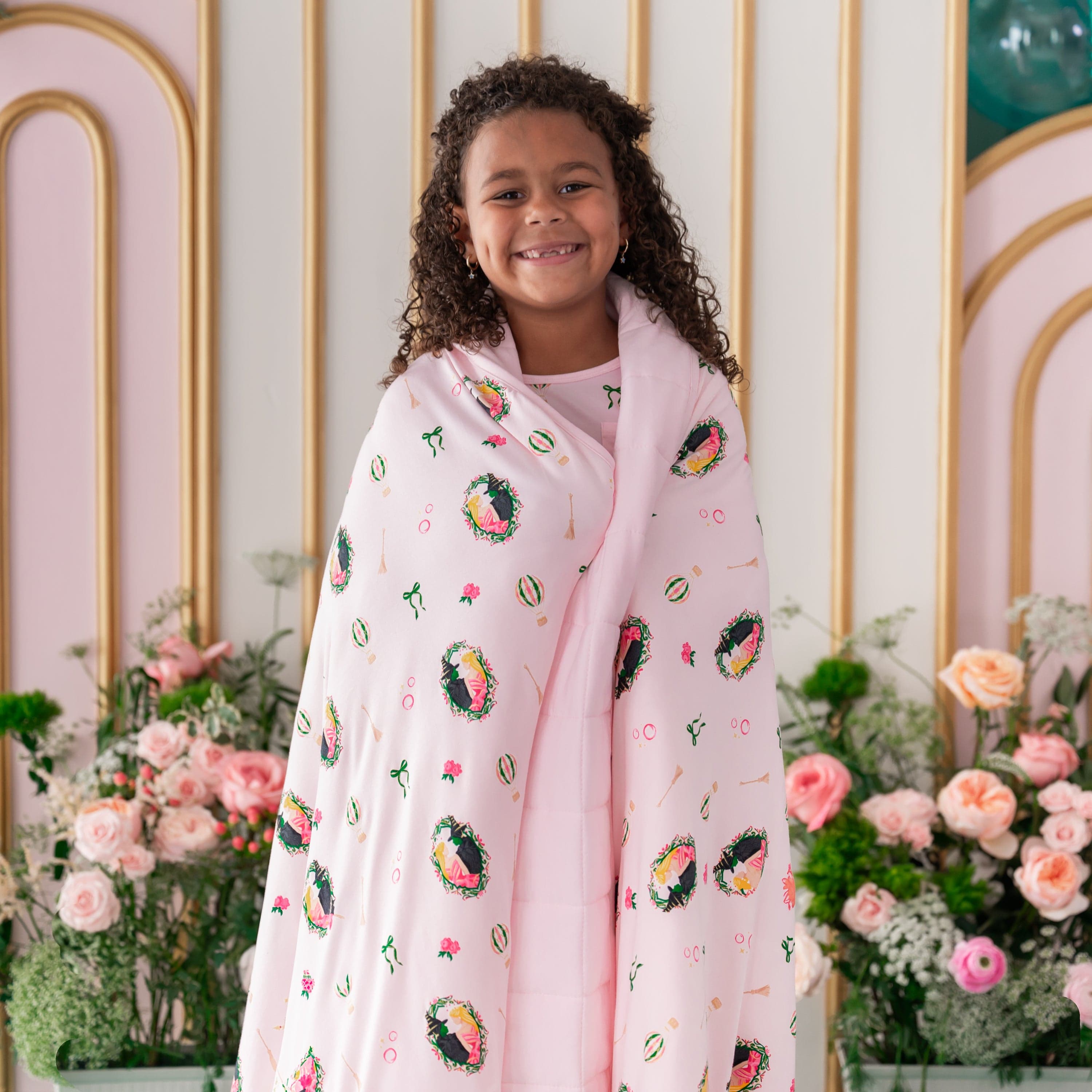Smiling girl standing in front of a floral backdrop with the Youth Blanket in Wicked 2.5 wrapped around her shoulders