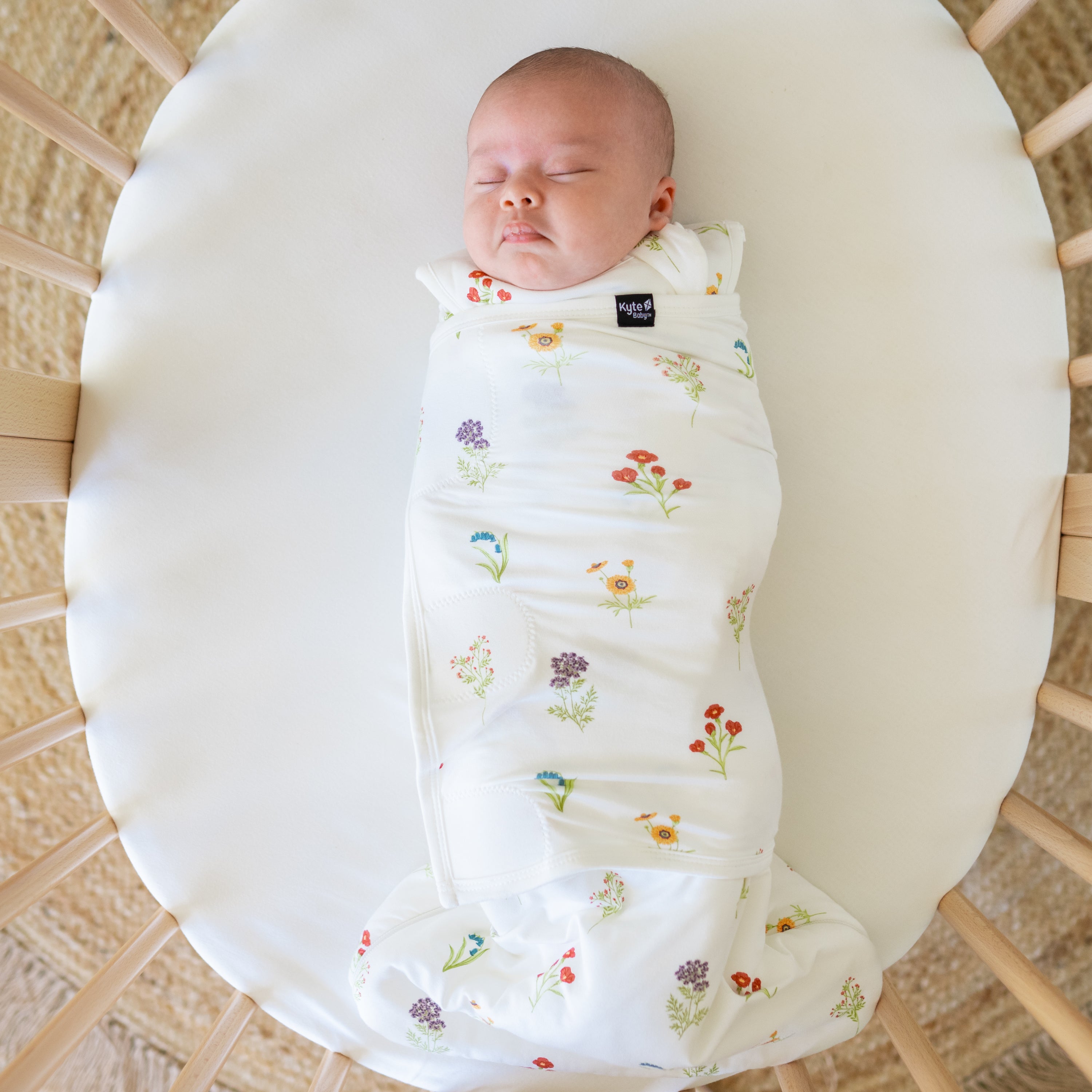 Close up of baby sleeping in a oval bassinet wearing Sleep Bag Swaddler in Wildflower 1.0