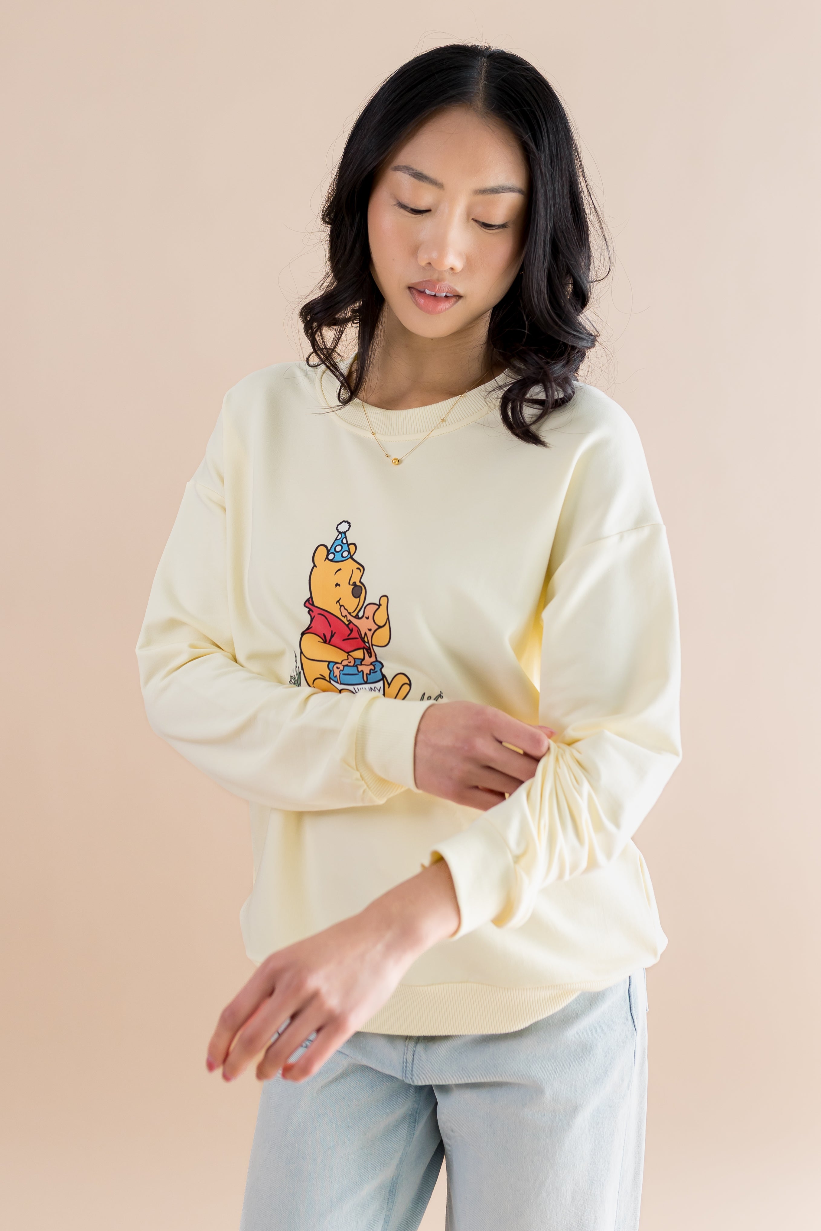 Female model wearing the Women's Sweatshirt in Winnie the Pooh on Chamomile rolling up the sleeves on the arm of the sweatshirt