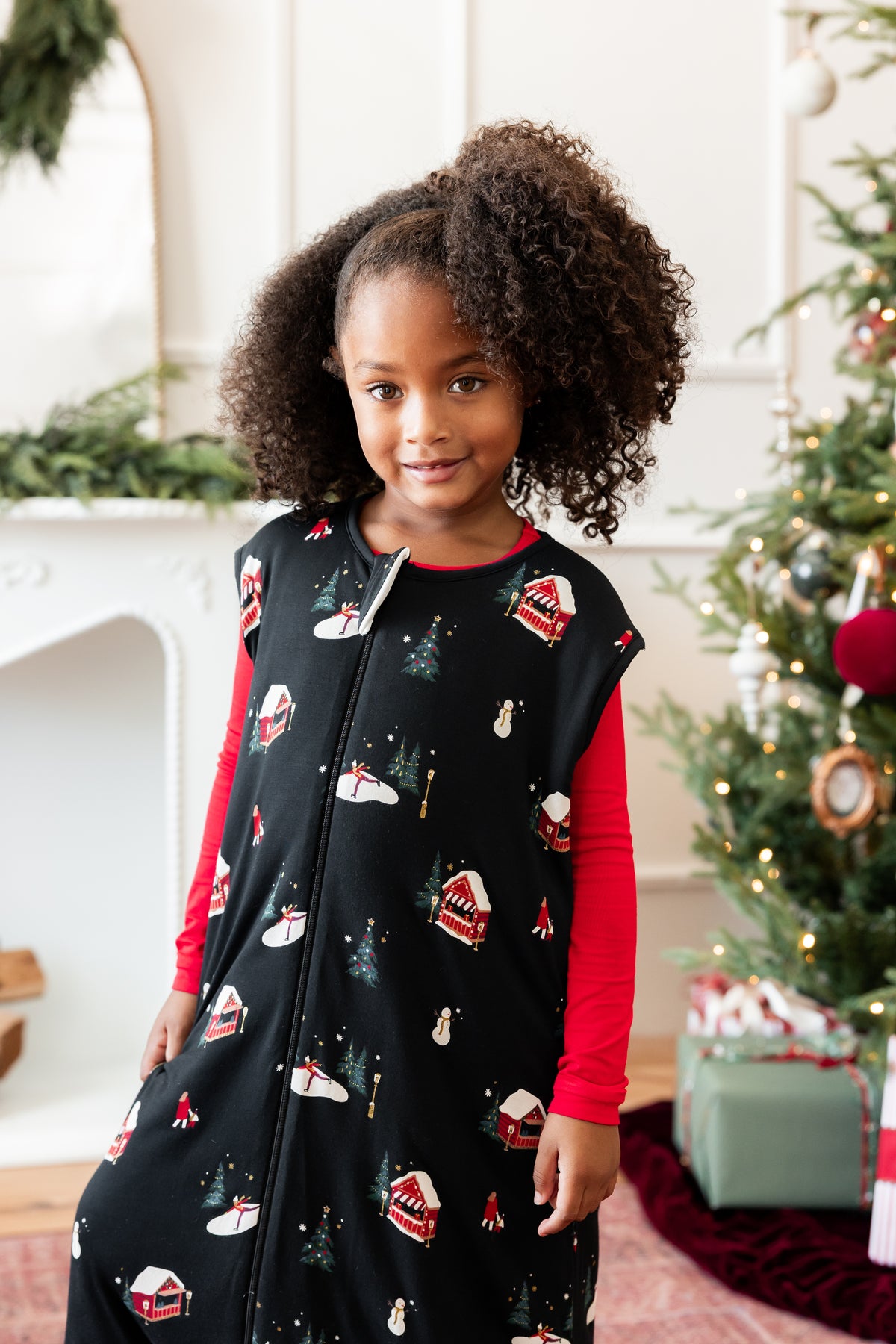 A young girl with curly brown hair stands in a holiday-themed room wearing a black zip-up sleep sack with a winter village print, layered over a red long-sleeve shirt. A decorated Christmas tree and fireplace are visible in the background.