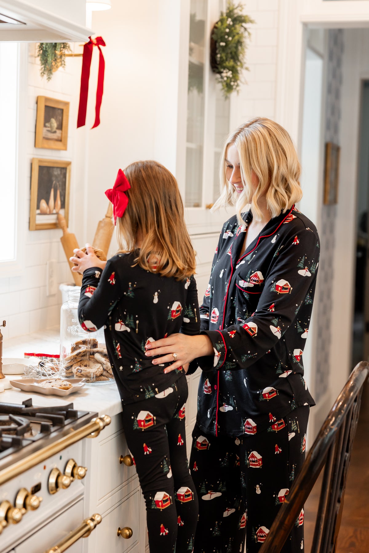 A woman and a young girl wearing matching black Christmas-themed pajamas with a holiday cookie print, standing in a bright kitchen. The pajamas feature red trim and red bow accents, and the girl has a red bow in her hair.