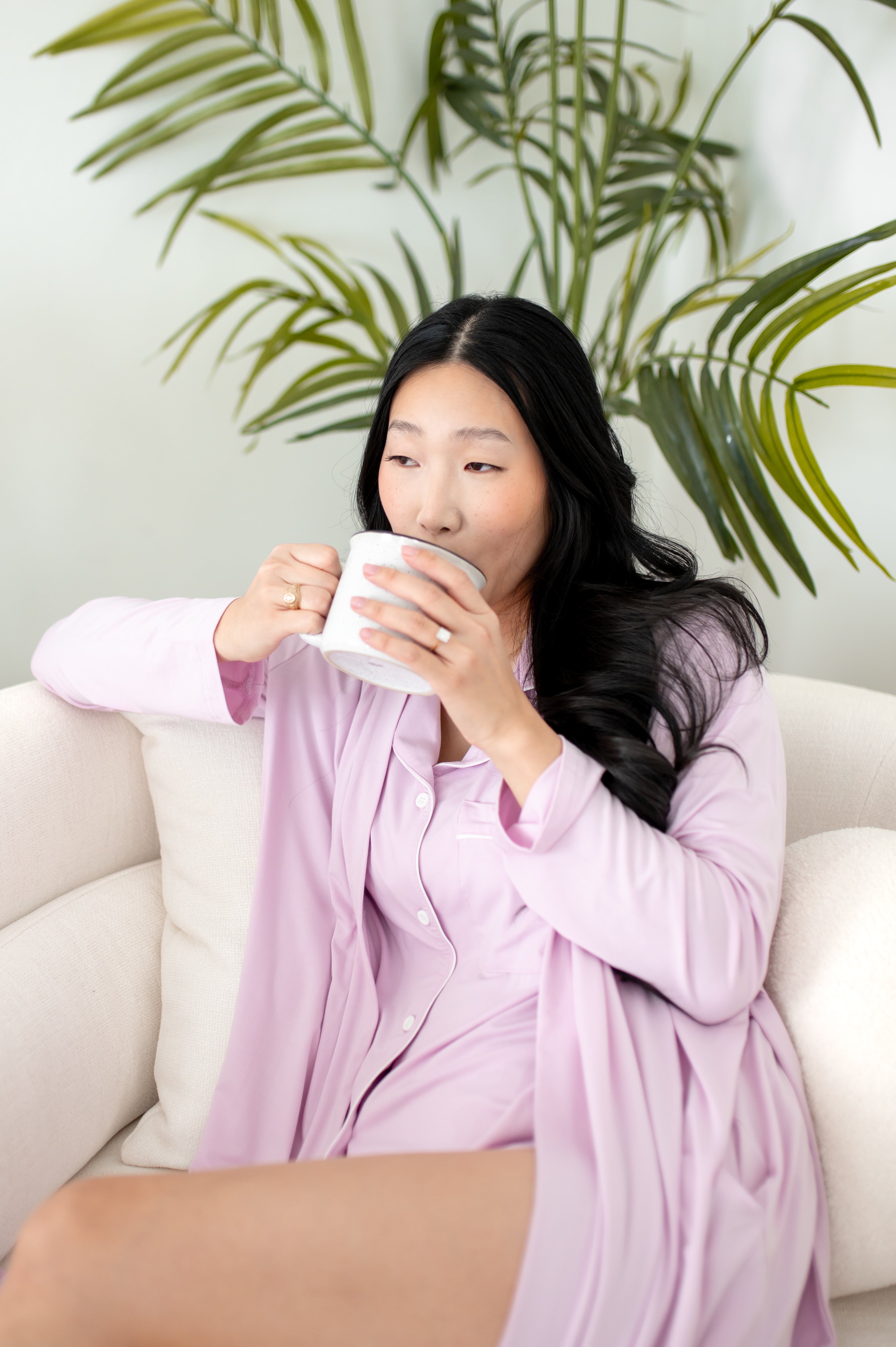 Woman in purple pajamas sitting on a white couch holding a mug with green plants in the background.