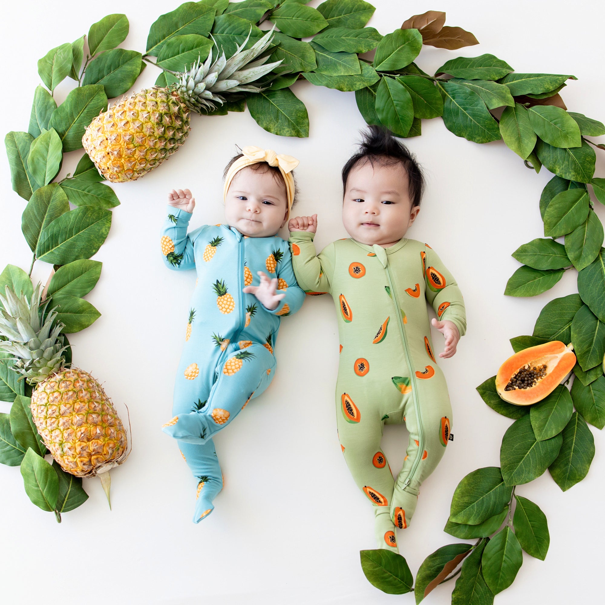 2 babies in zippered footies in prints papaya and pineapple surrounded by fruit and leaves