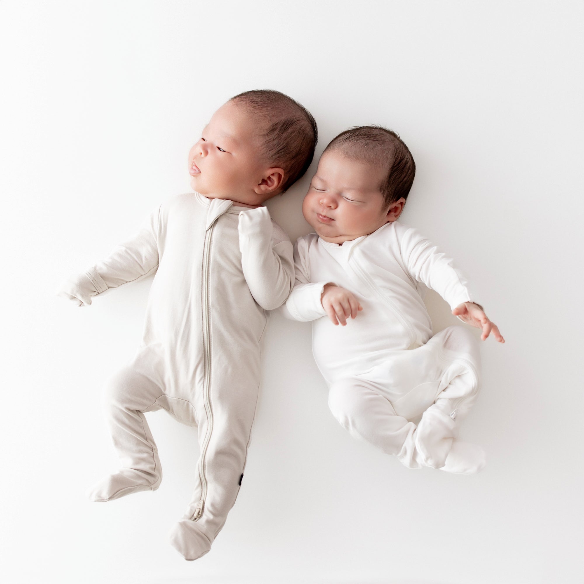 Two infants laying next to each other while wearing Kyte Baby Zippered Footies in Oat and Cloud