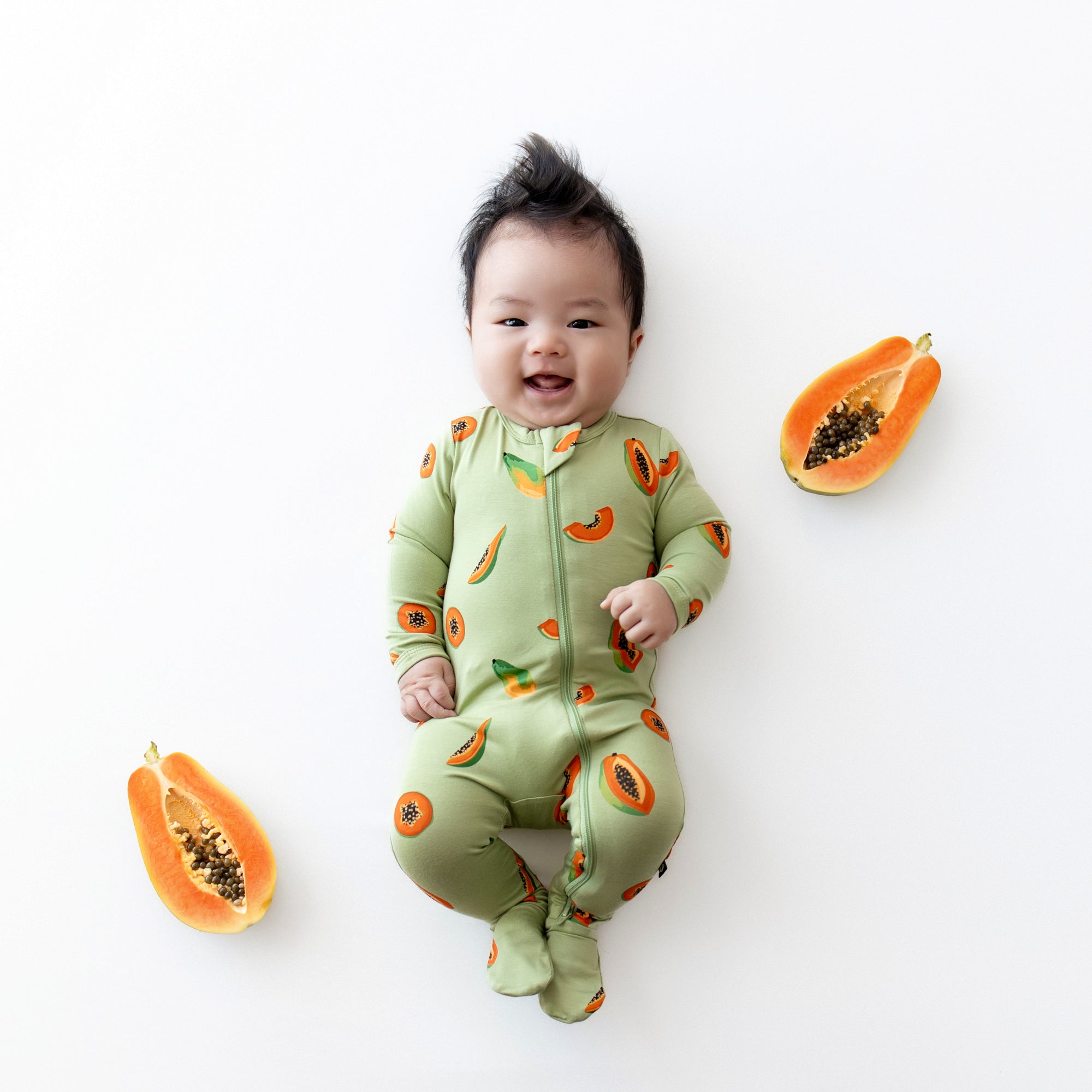 Baby in zippered footie in papaya laying next to real papayas