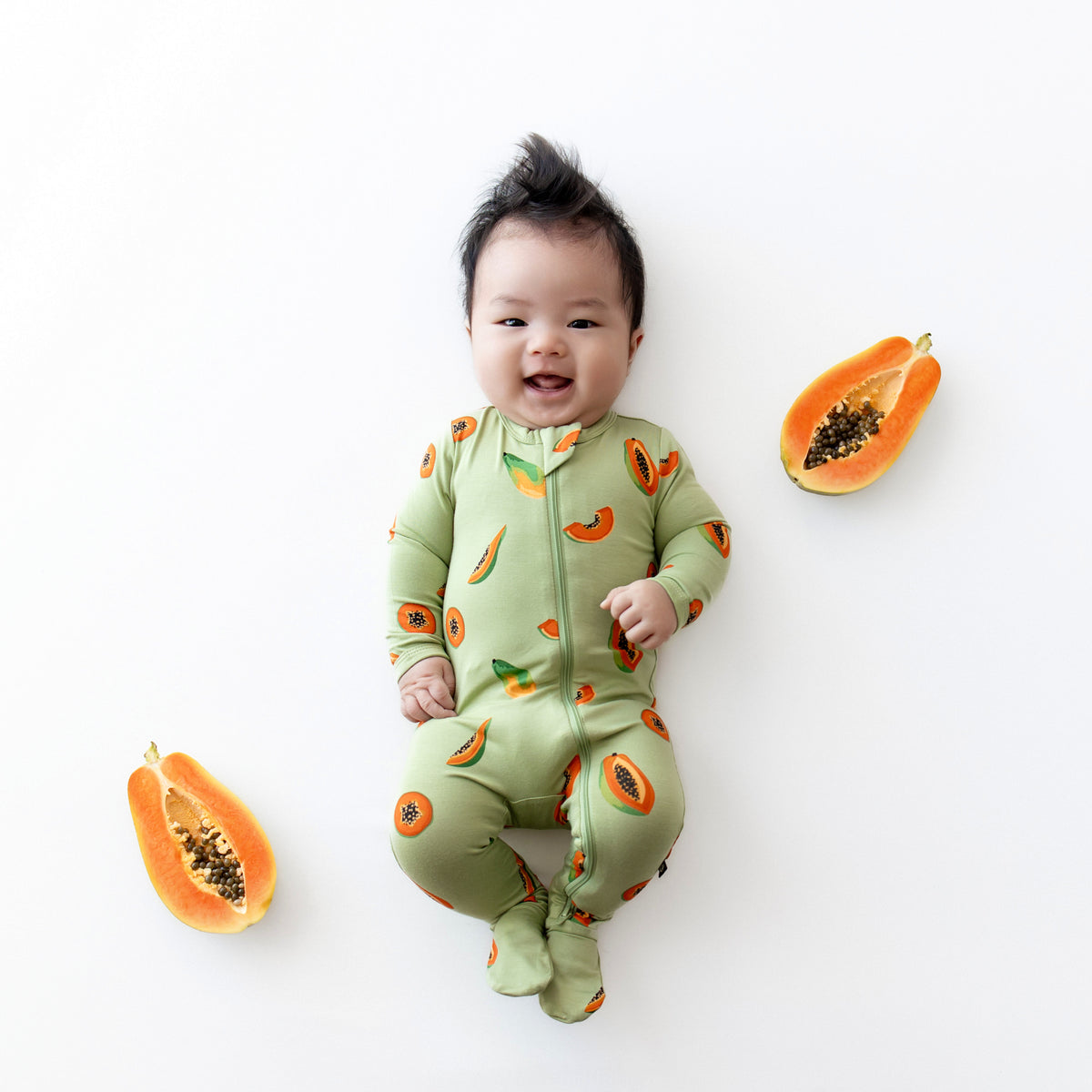 Baby in zippered footie in papaya laying next to real papayas