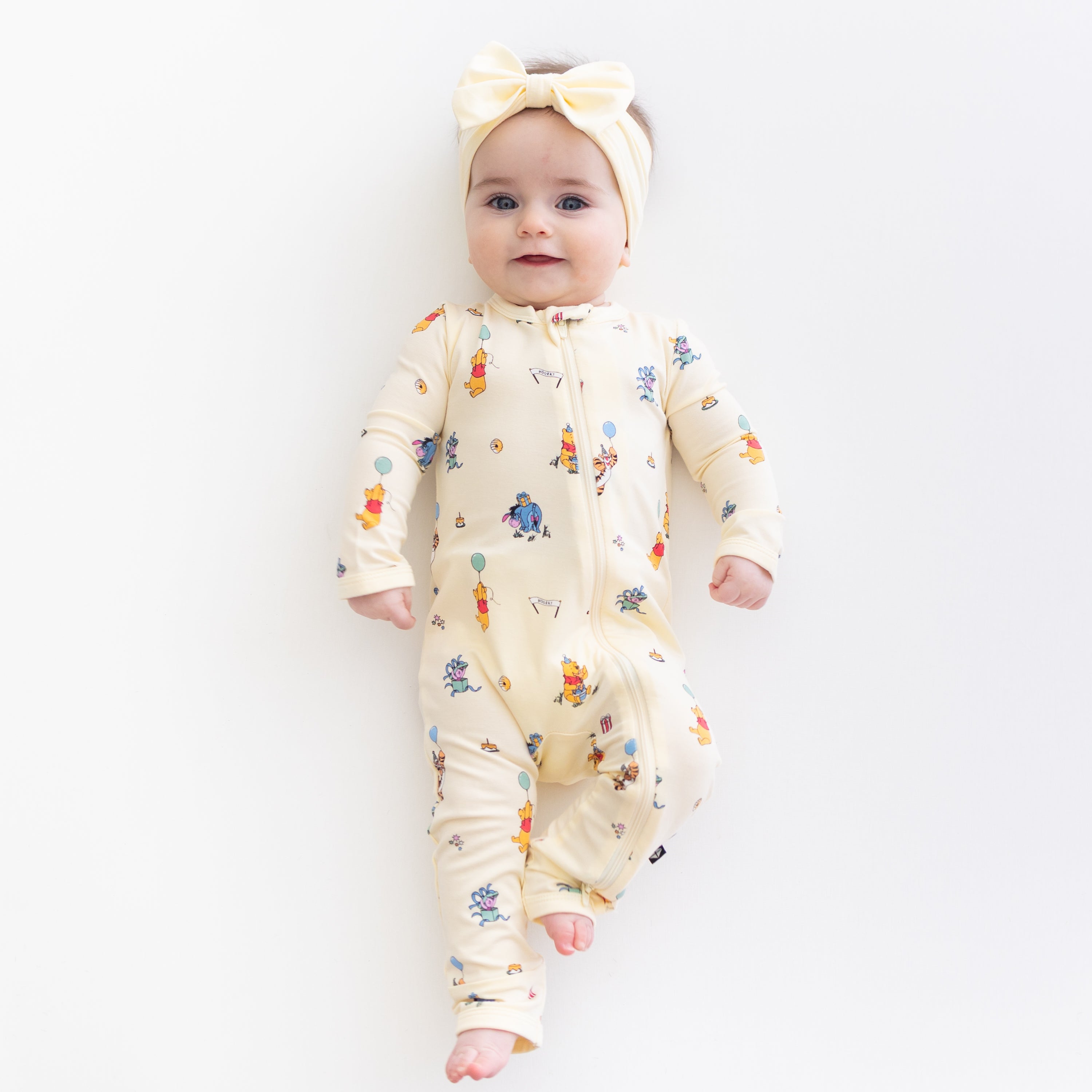 Young toddler laying on a light neutral surface wearing the Zippered Romper in Winnie the Pooh Celebration with a light yellow bow