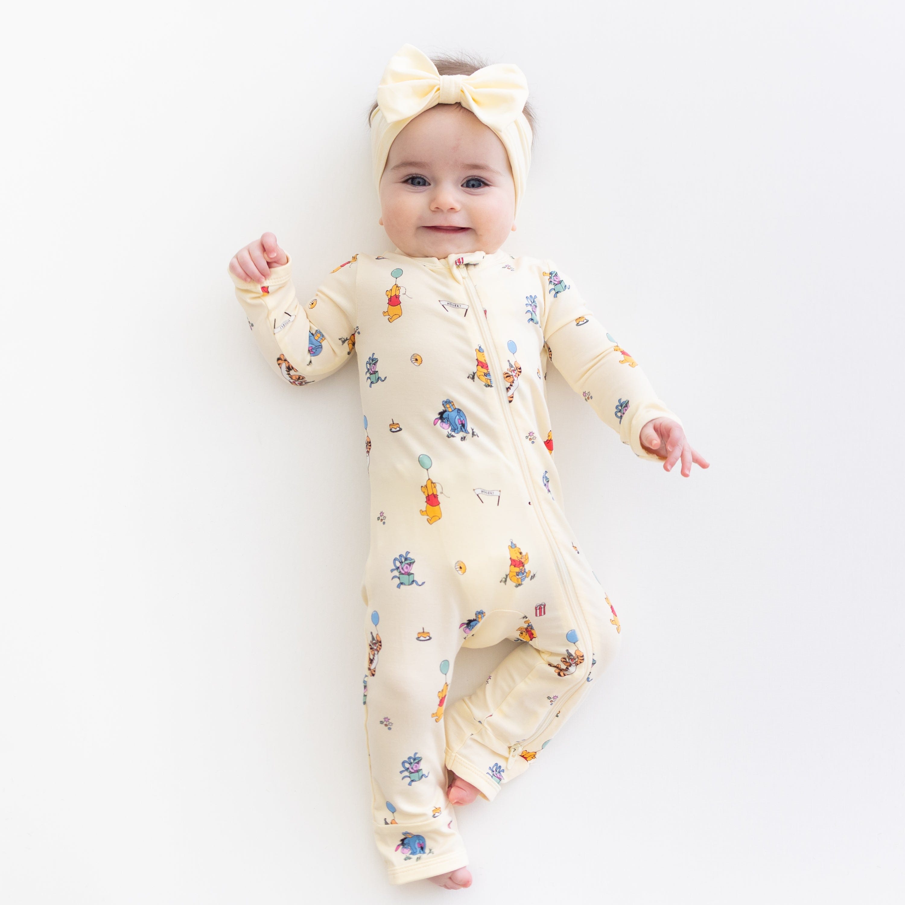 Young toddler laying on a light neutral surface wearing the Zippered Romper in Winnie the Pooh Celebration with a light yellow bow