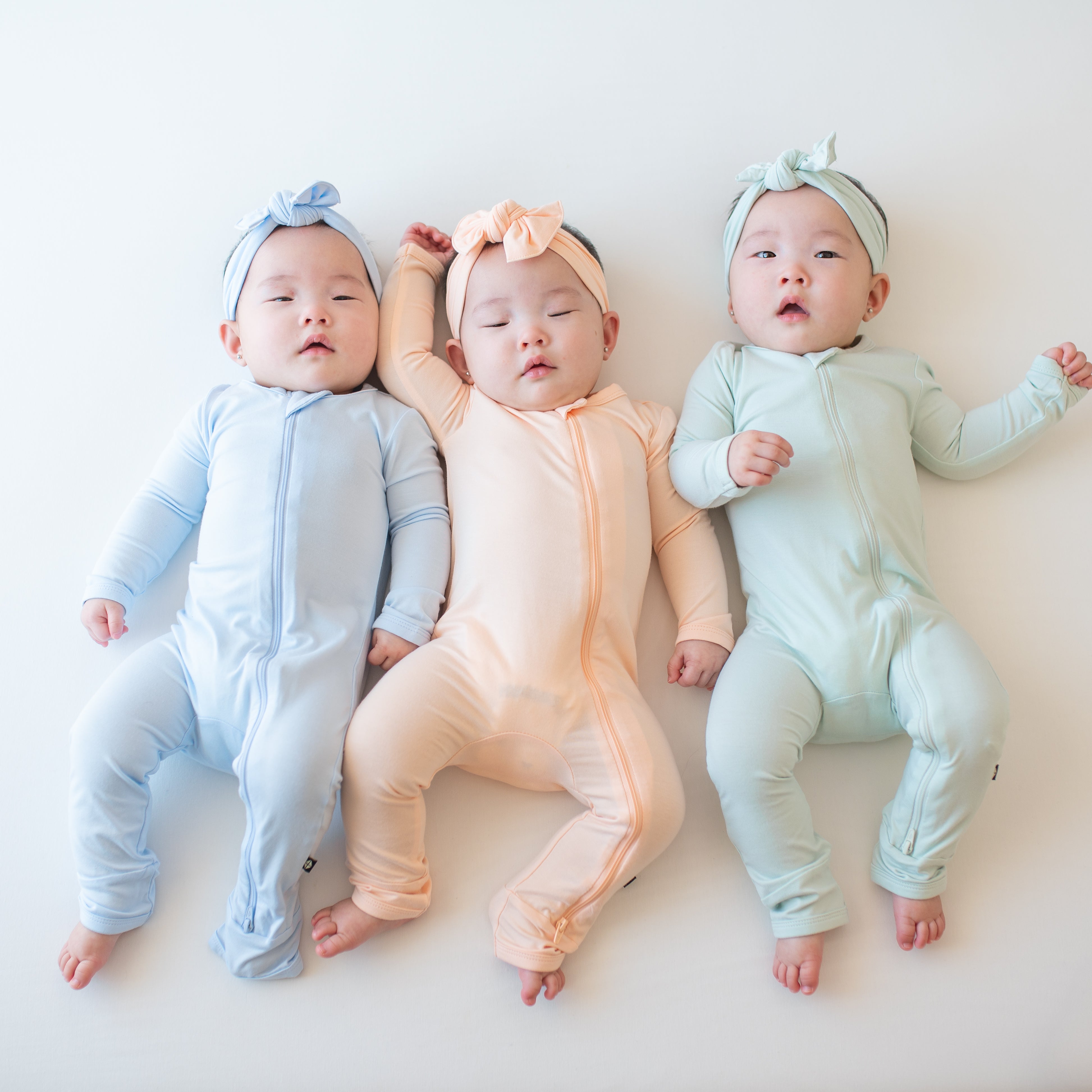 Three infant models wearing zippered rompers and matching bows in the Summer solid colors